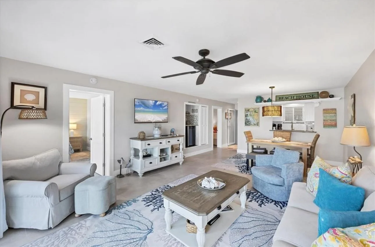 Coastal-themed living room with white couches, a blue armchair, floral pillows, a wooden coffee table, a white console with a TV, a round dining table with chairs, beach decor, and a ceiling fan.