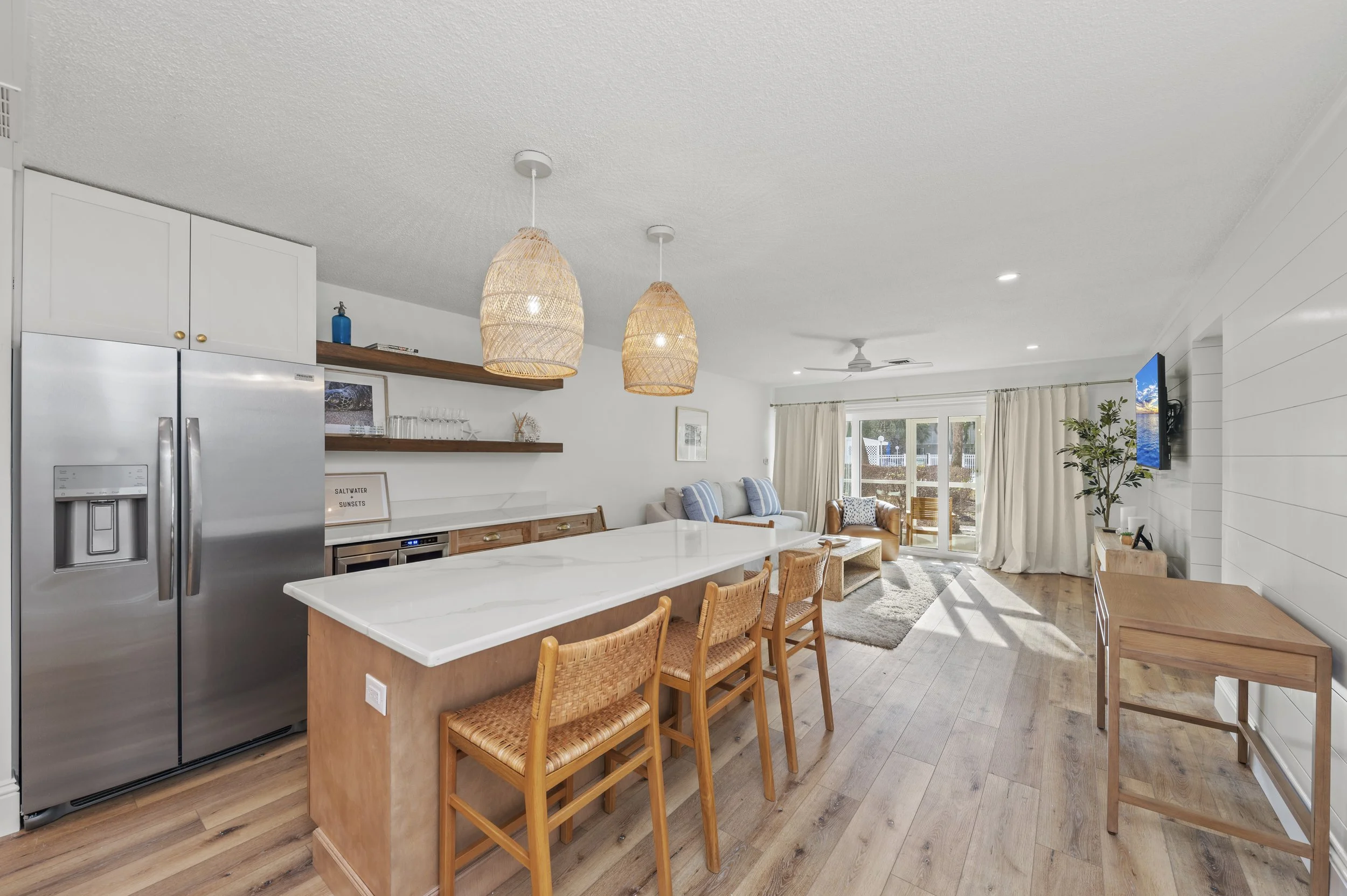 Open-concept kitchen and living room with white walls, wooden flooring, and natural light from large sliding glass doors. The kitchen features a large island with a white countertop, wooden barstools with woven seats, stainless steel refrigerator, an