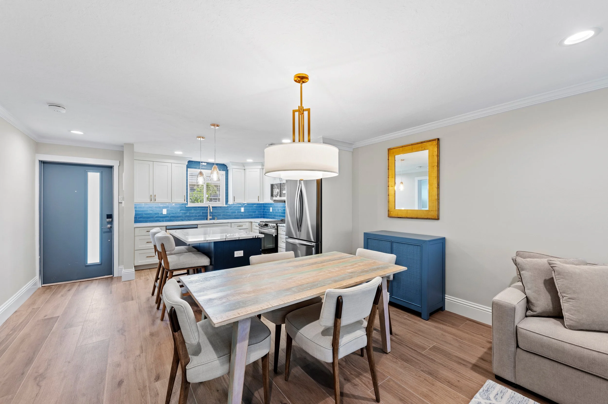 Open-concept kitchen and dining room with breakfast bar, wooden dining table, beige chairs, gray sofa, blue cabinet, yellow framed mirror, and modern chandelier.