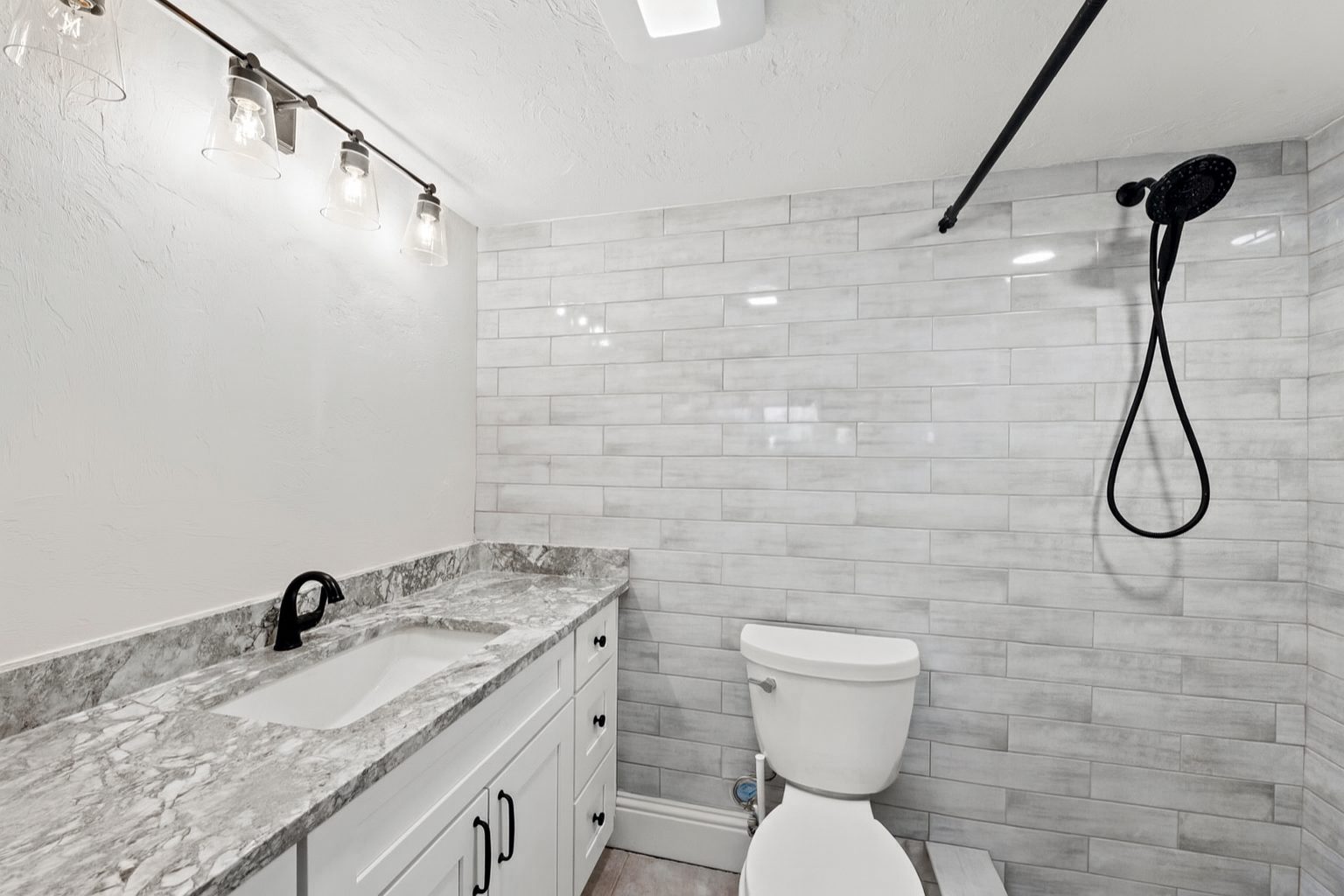 Bathroom with white vanity, marble countertop, black faucet, gray tiled walls, toilet, and black showerhead with a flexible hose.