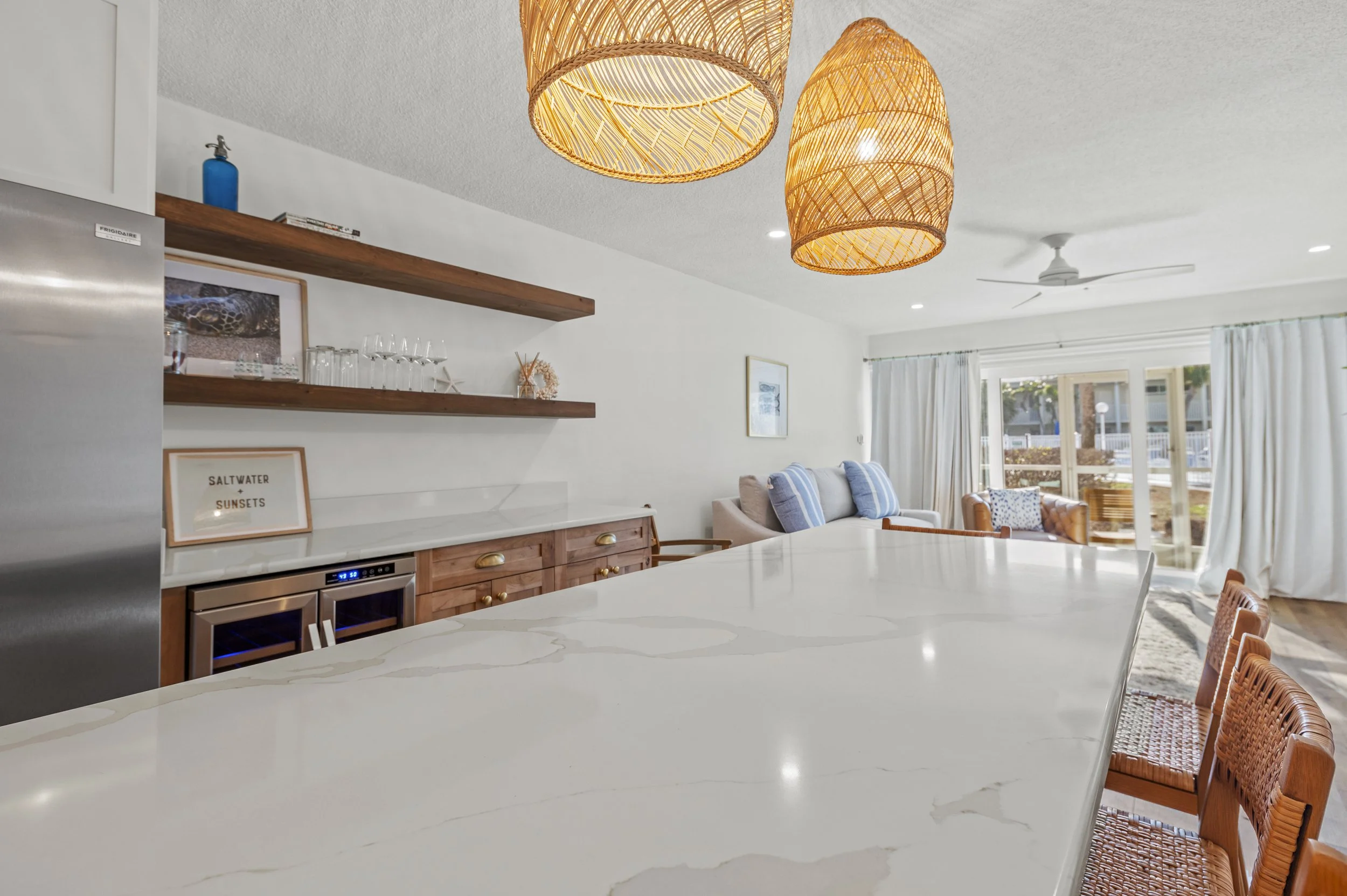 Open-concept kitchen and living room with white walls, wooden accents, hanging wicker light fixtures, a marble island, and large window with white curtains.