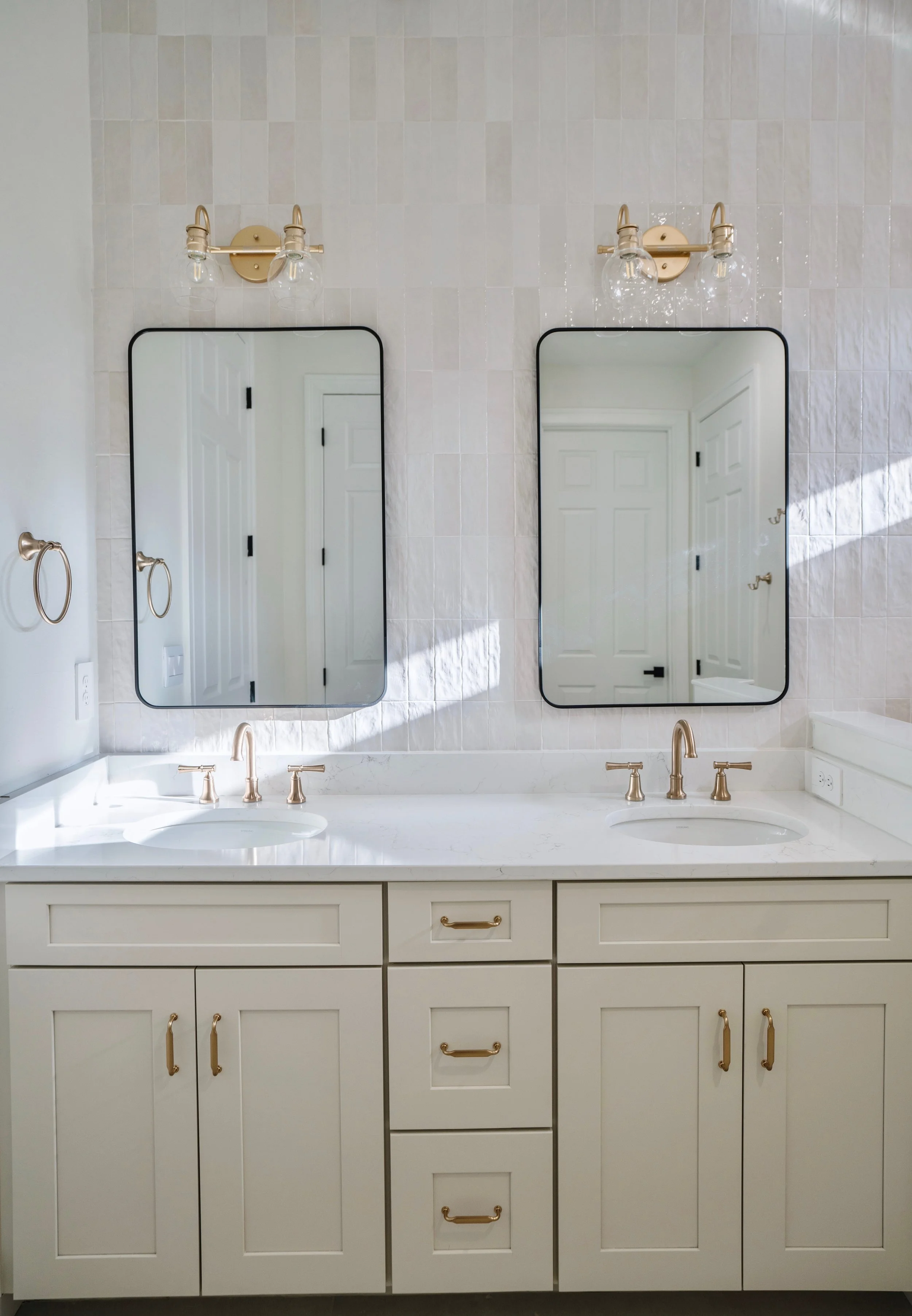 A bathroom double vanity with a white marble countertop, two oval sinks, and gold fixtures. Two rectangular mirrors with black frames hang above each sink, with wall-mounted gold light fixtures above each mirror. The wall behind the vanity is tiled w