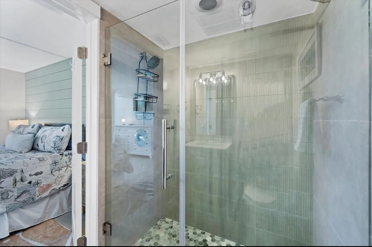 A glass shower enclosure with green tiled walls and a hexagonal tile floor in a bathroom. Inside the shower, there is a showerhead, a soap dispenser, and a metal shelf. Reflected in the mirror, there is a glimpse of a bathroom vanity and a chandelier