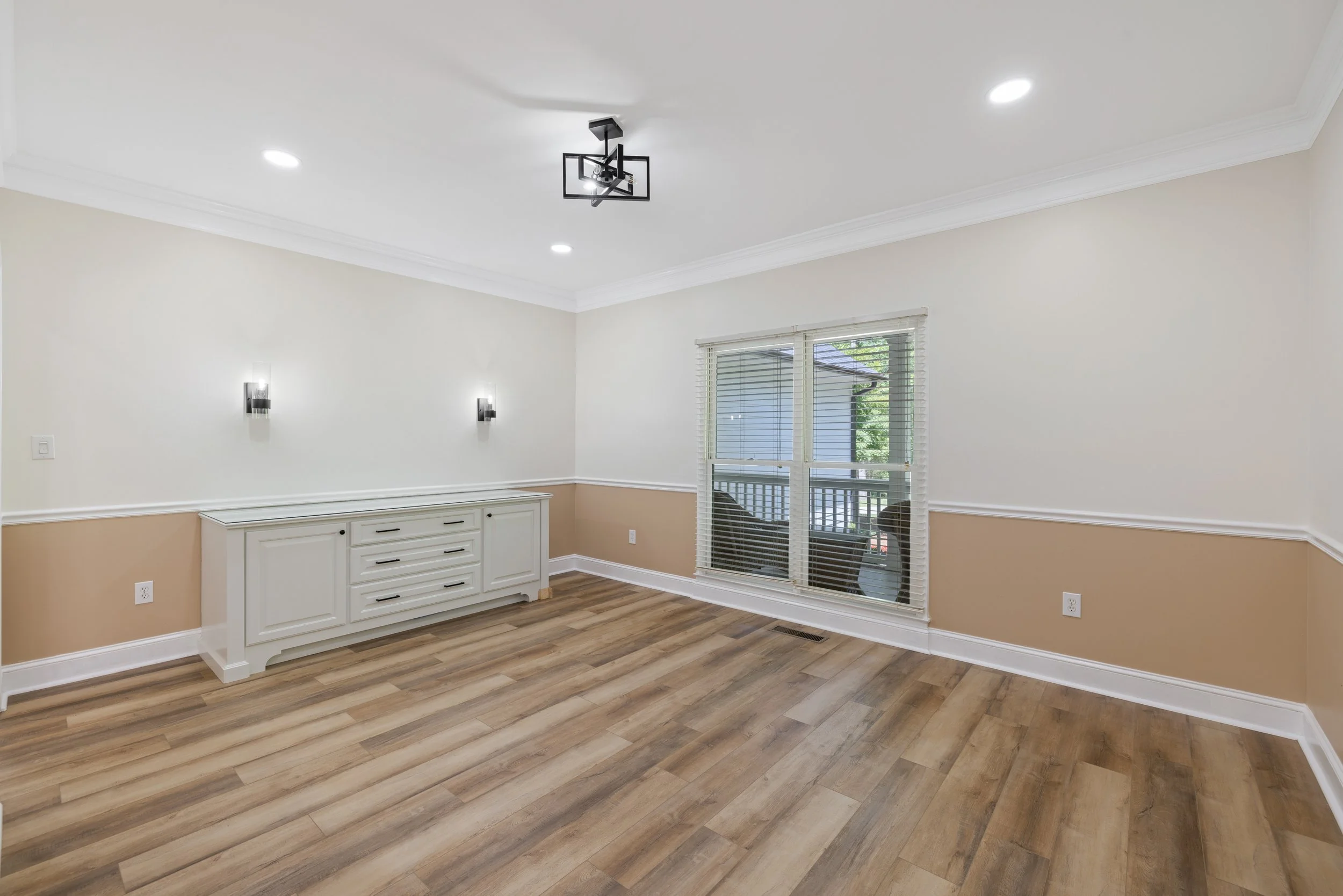 Empty dining room with hardwood floors, white walls with beige wainscoting, a white sideboard, two wall sconces, a window with blinds, and a modern ceiling light fixture.