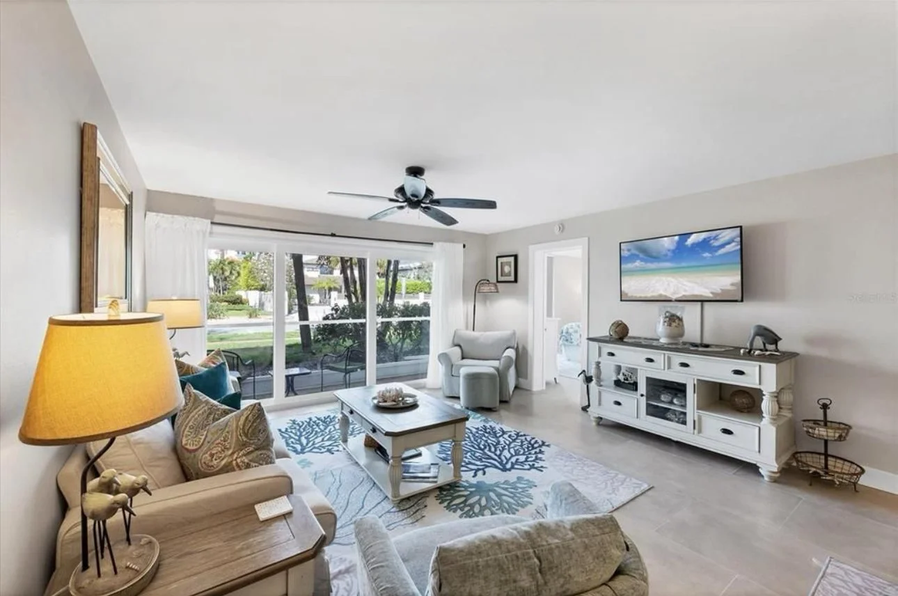 Bright living room with white walls, a white sofa with patterned pillows, a small wooden table, a white armchair, a white entertainment console with a flat-screen TV depicting a beach scene, and large sliding glass doors with white curtains leading t