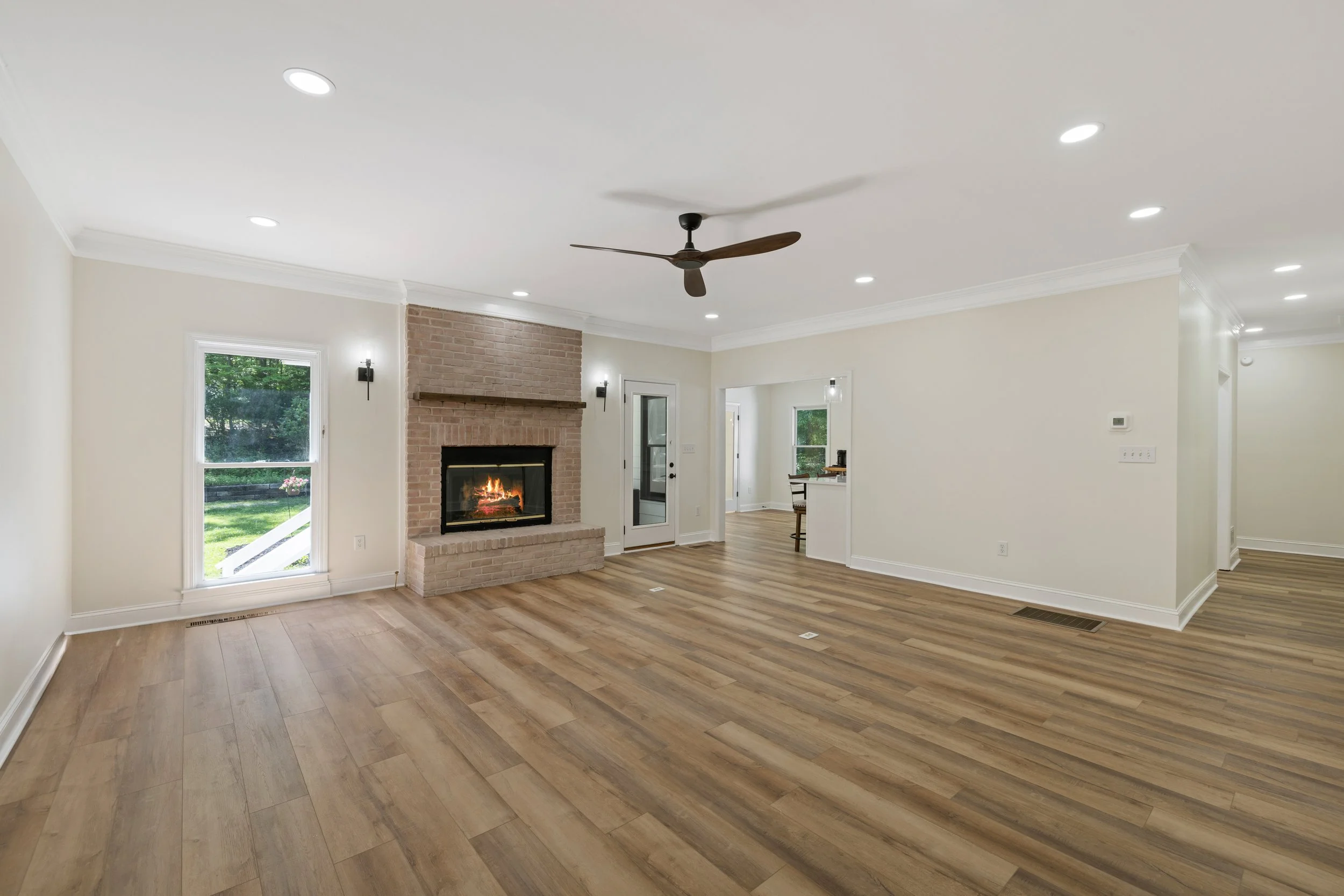 Empty living room with hardwood floors, a brick fireplace, ceiling fan, multiple recessed lights, and windows showing a yard outside.