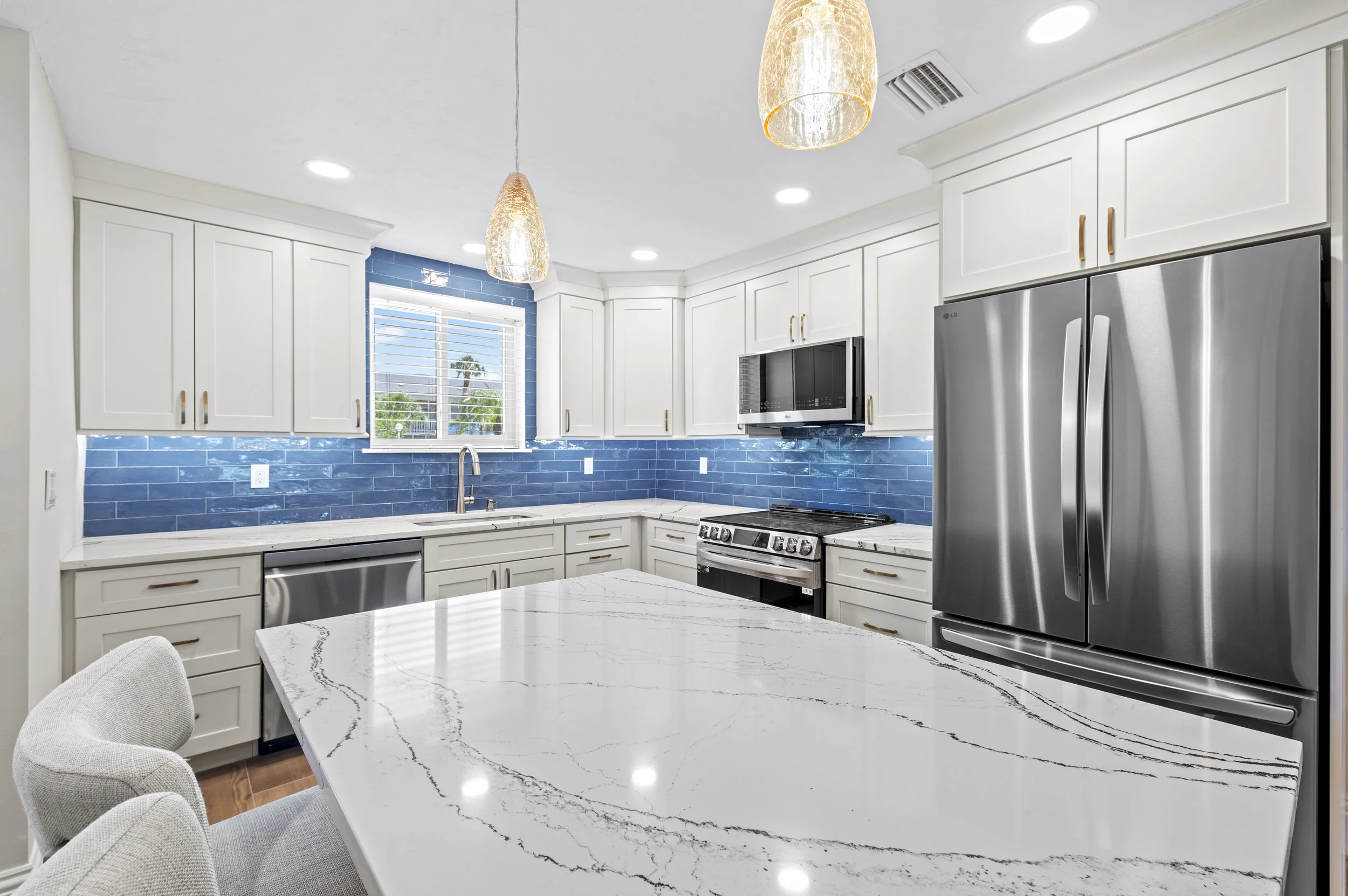 Modern kitchen with white cabinets, blue tile backsplash, marble countertop island, stainless steel appliances including a refrigerator and oven, window with blinds, and pendant lighting.