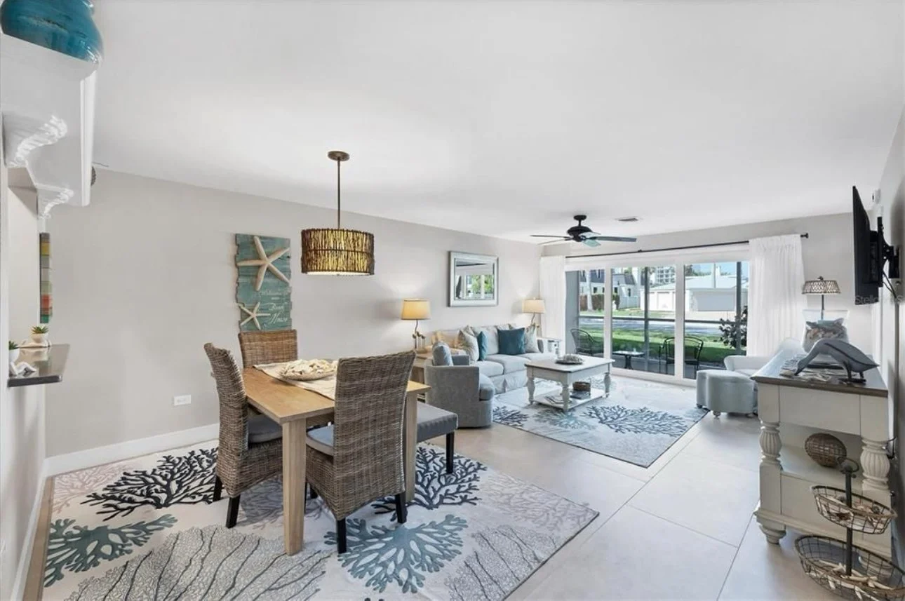 Living room and dining area with white walls, large sliding glass door, beige tiled floor, and coastal decor including starfish wall art. The room has a dining table with four chairs, a white sofa with blue throw pillows, a white armchair, and a whit