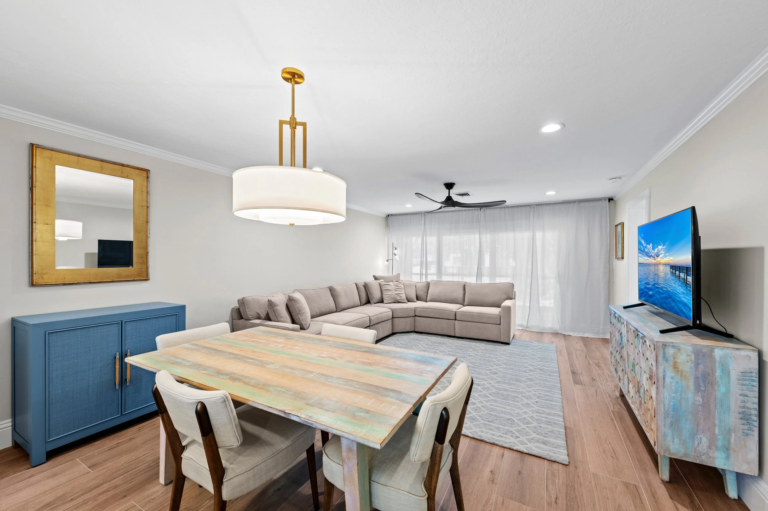 Living room with beige sectional sofa, wooden dining table with five chairs, blue sideboard, wall mirror, flat-screen TV on a colorful console, ceiling fan, and sheer curtains.
