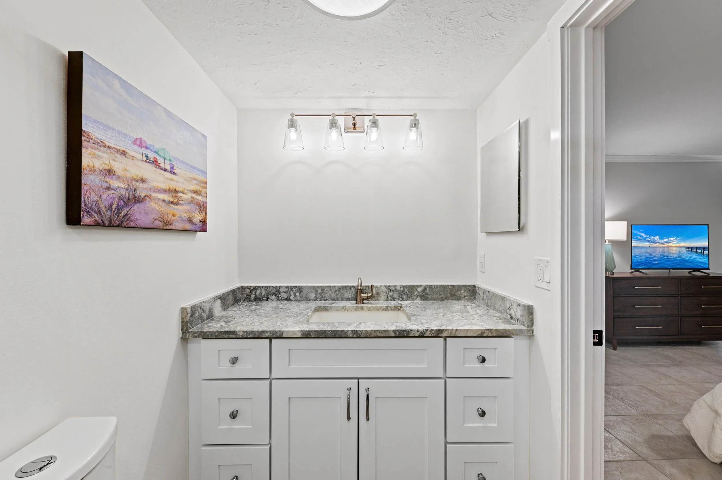 Bathroom vanity with a marble countertop, white cabinets, a small sink, and a bronze faucet. There is a wall-mounted light fixture with four clear glass shades above the vanity. A colorful beach scene painting hangs on the left wall. Part of a toilet