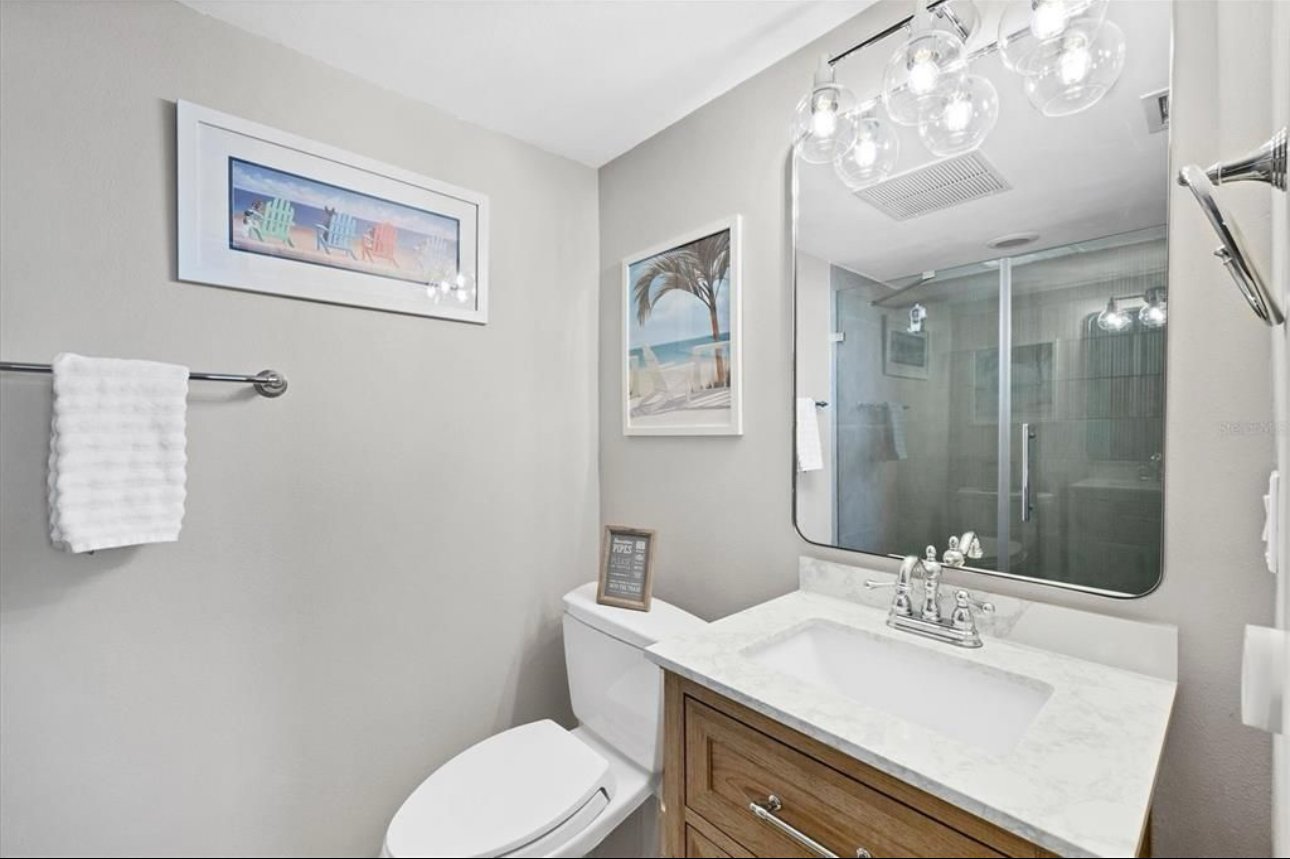 A small bathroom with a white toilet, a wooden vanity with a white marble countertop and an integrated sink, a large mirror above the sink, a framed picture of a beach scene on the wall, a towel bar with a white towel, and wall art depicting colorful