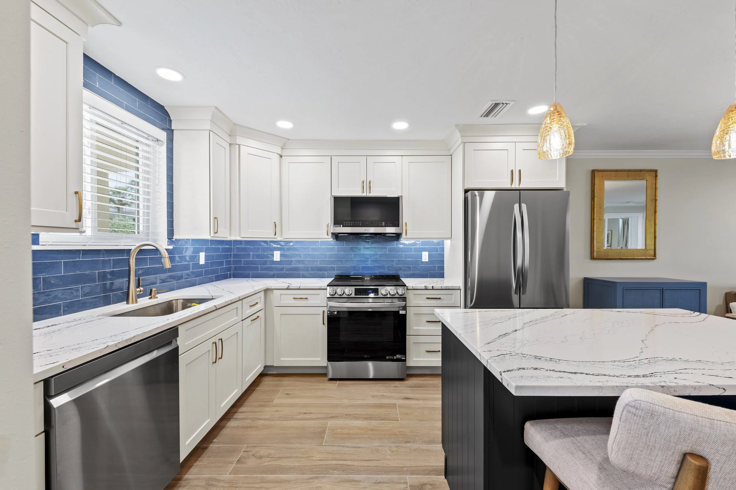 Modern kitchen with white cabinets, blue tiled backsplash, marble countertop, and stainless steel appliances including refrigerator, stove, and microwave. There is an island with a marble top and a beige chair, with a wooden floor and a blue side tab