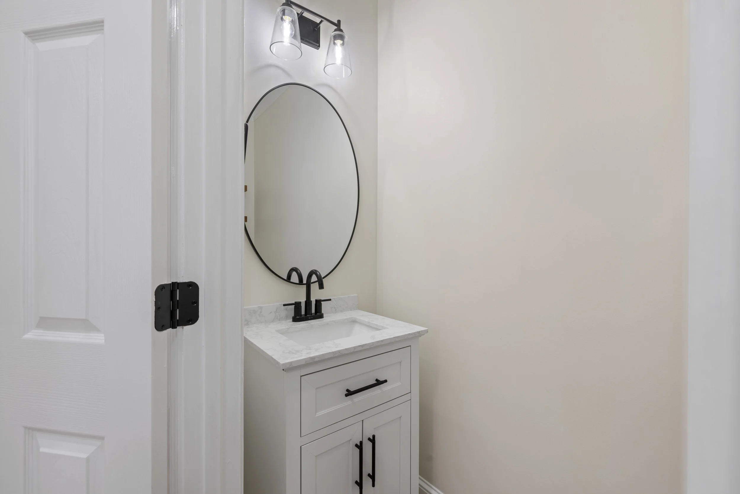 A small bathroom vanity with a white cabinet, black handles, a white marble countertop, black faucet, oval mirror, and modern light fixture with two glass shades.