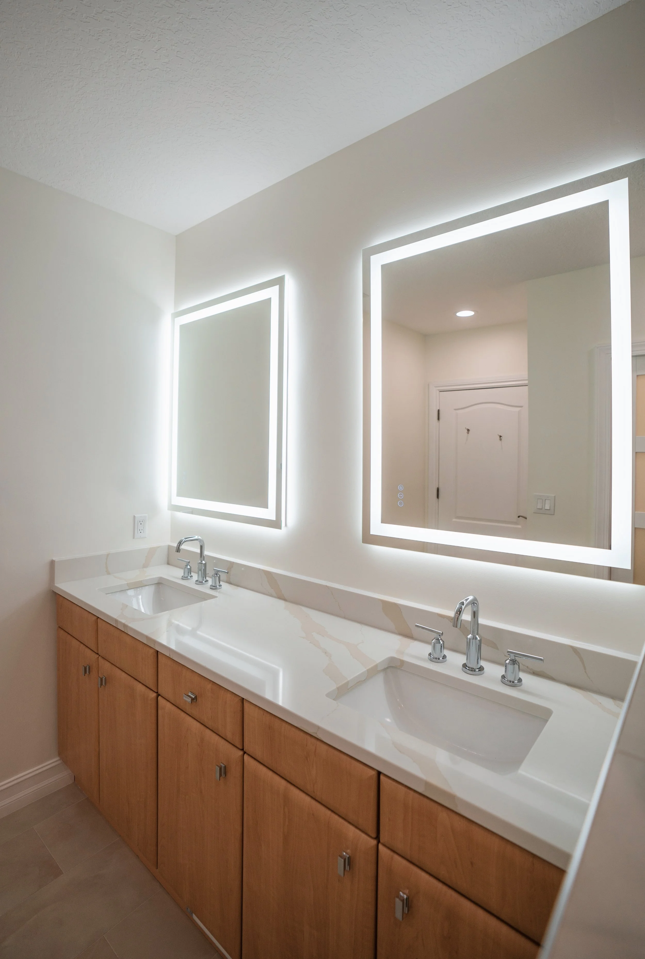 Modern bathroom with wooden vanity, white marble countertop, double sinks, illuminated mirrors, and a door reflected in the mirror.