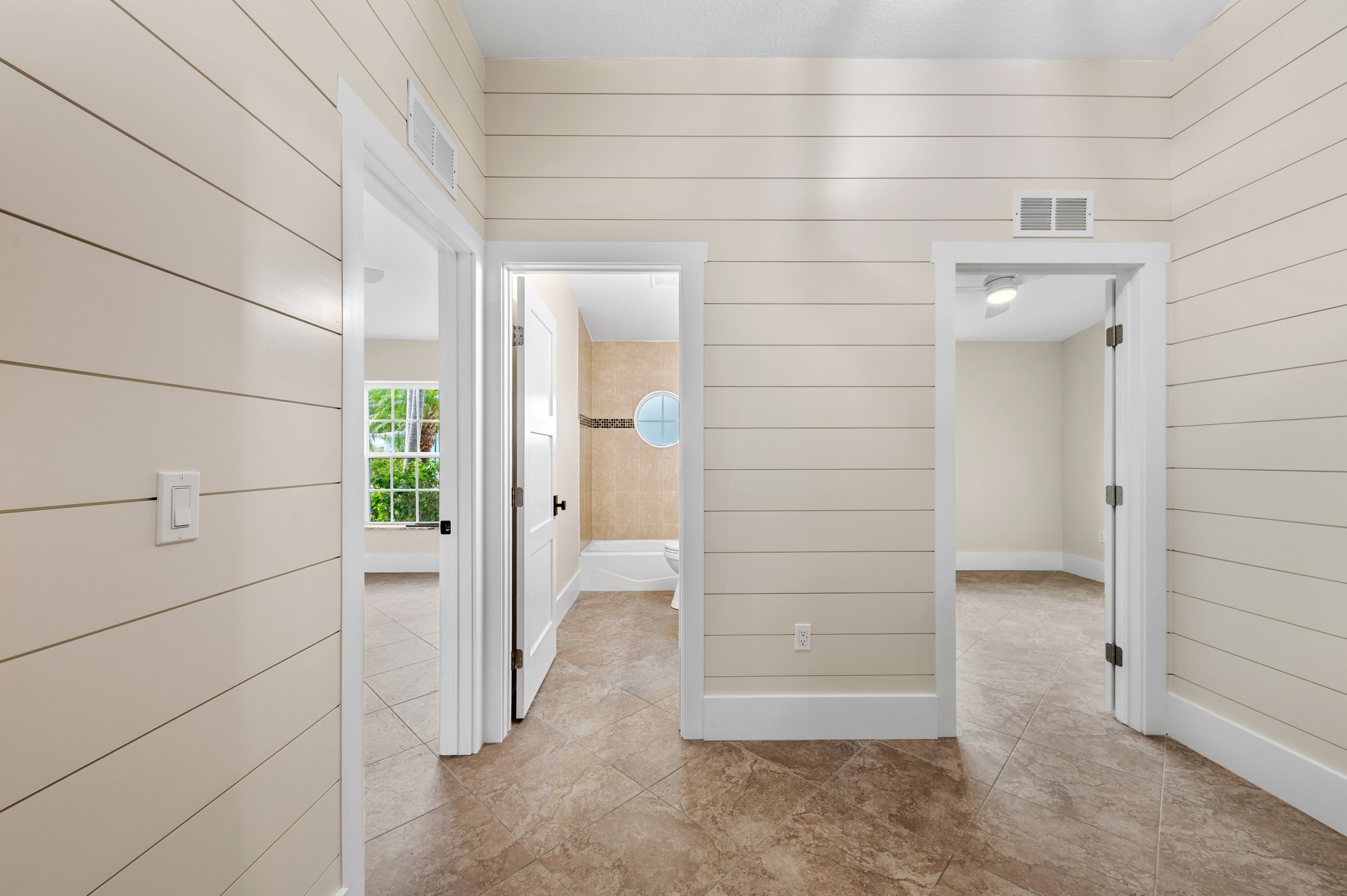 Interior of a house with tiled floors, beige walls with horizontal paneling, and doorways leading to other rooms including a bathroom with a circular window.