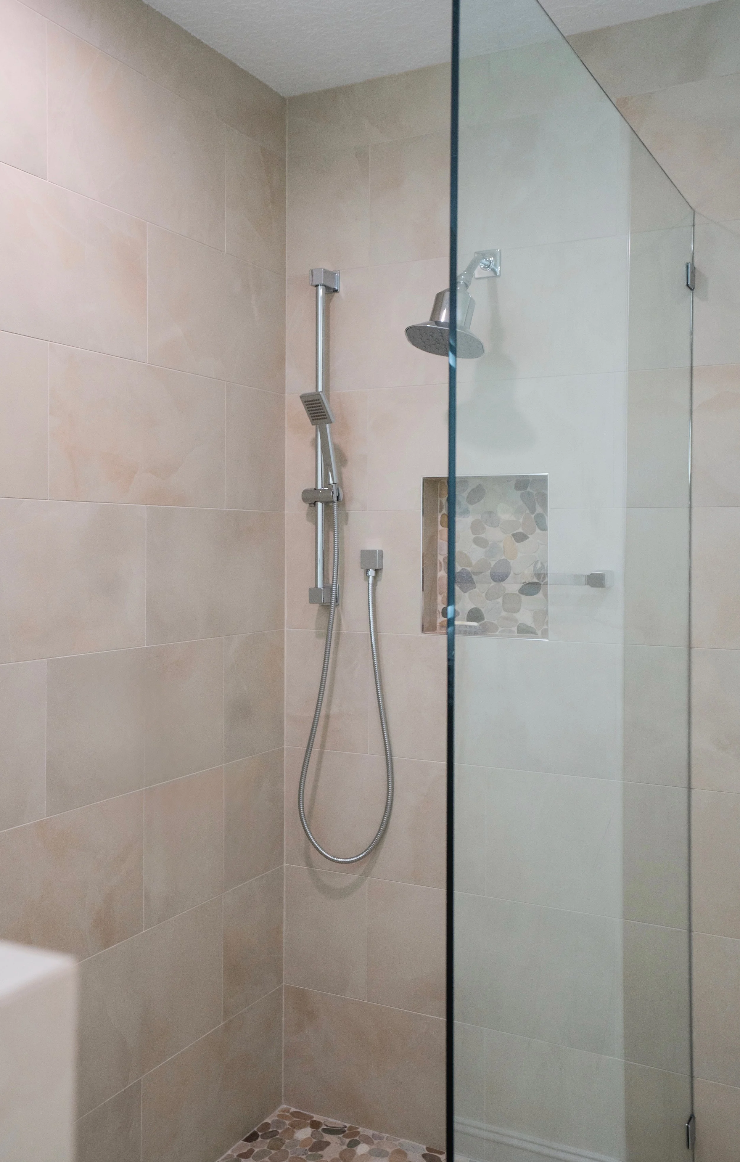 A walk-in shower with beige tile walls, a rainfall showerhead, a handheld showerhead, and a pebble tile floor, enclosed by a glass panel.