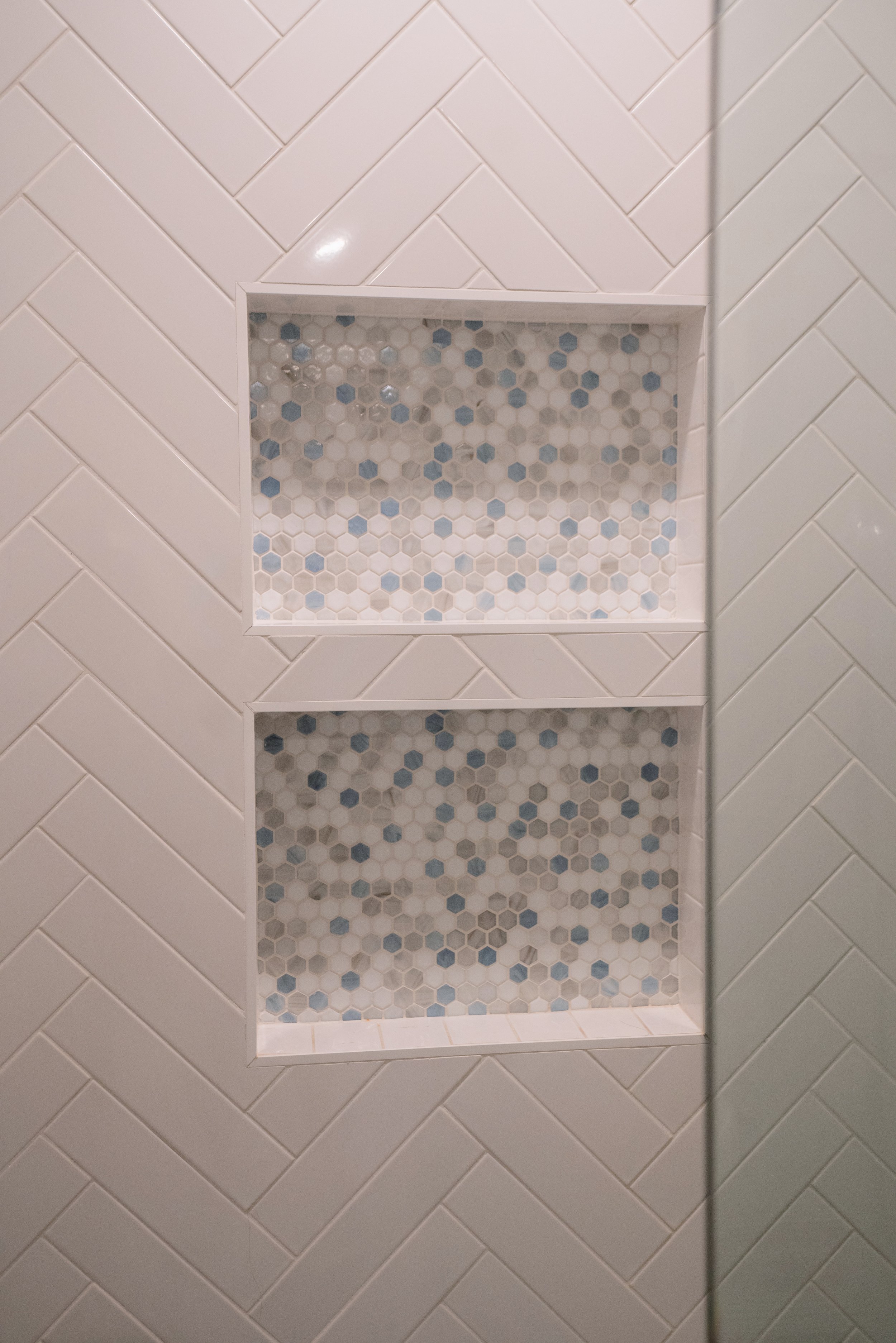 Bathroom shower wall with white herringbone tiles and two built-in tiled shelves with hexagonal tiles in shades of blue, gray, and white.