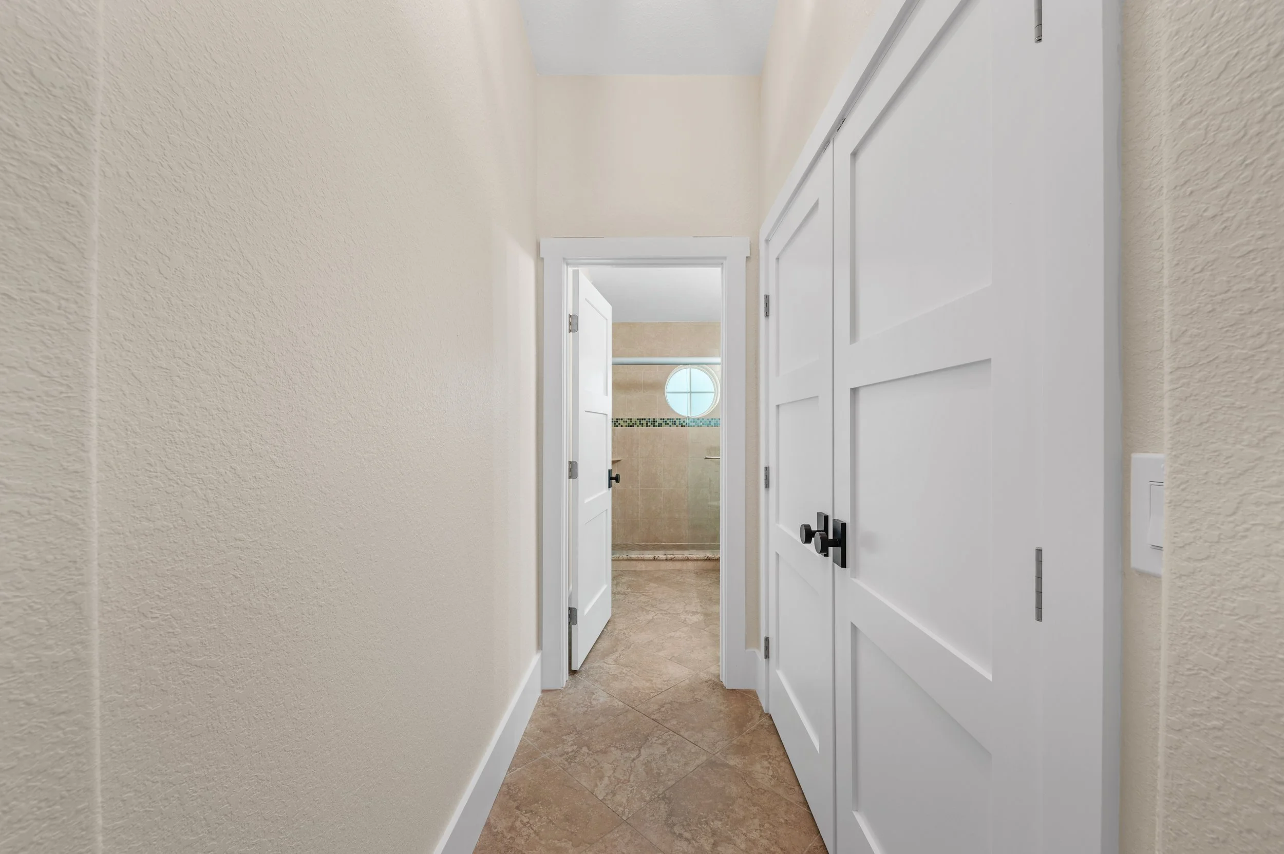 A narrow hallway with beige walls, white trim, and a tiled floor leading to a bathroom with a round window and beige tiles.