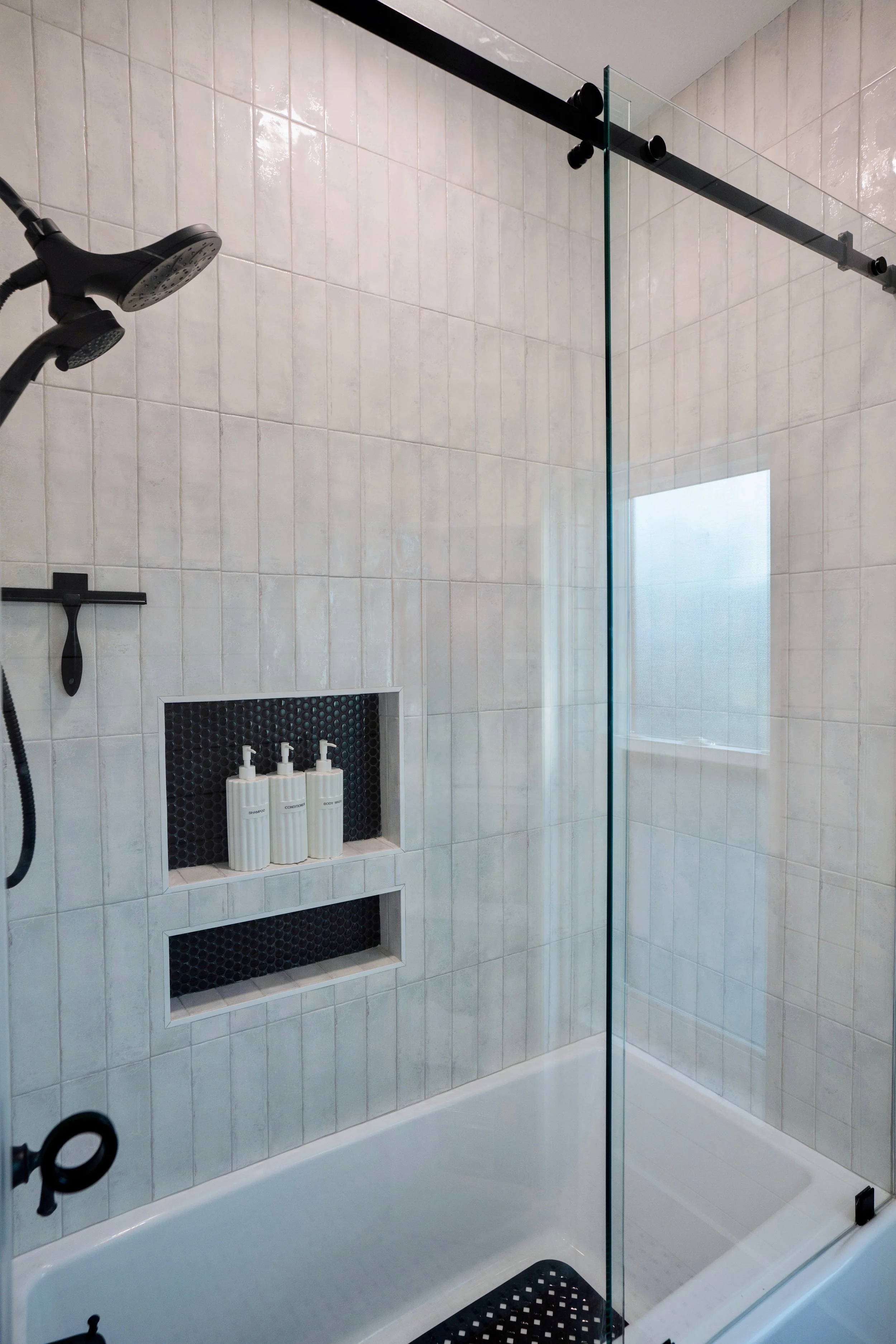A modern shower with beige tiled walls, a glass partition, and built-in niches for toiletries. There is a black showerhead and soap dispensers in the niches.