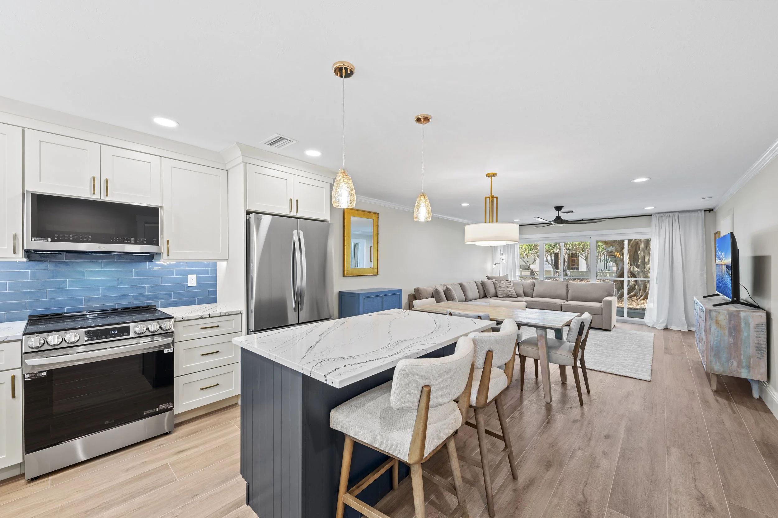 Open-concept kitchen and living room with white cabinetry, stainless steel appliances, a blue tile backsplash, a kitchen island with white marble countertop, and a living area with a large sectional sofa and a flat-screen TV.