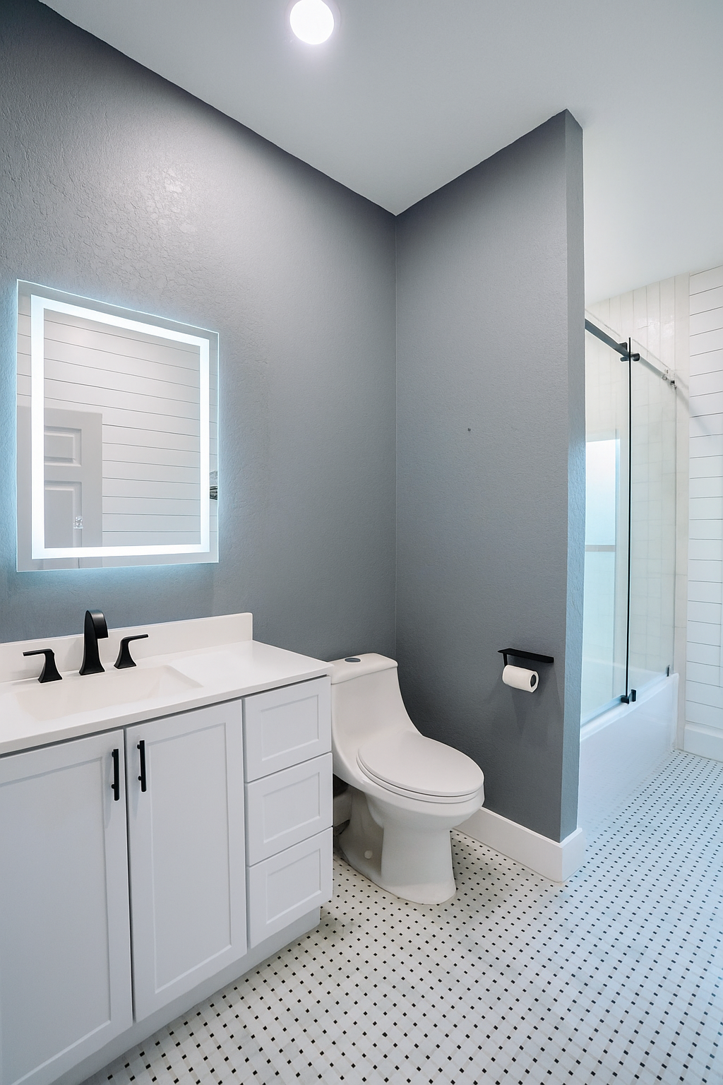 Modern bathroom featuring a white vanity with black fixtures, a wall-mounted mirror with LED lighting, a white toilet, a toilet paper holder, and a shower with glass door and white tiles.