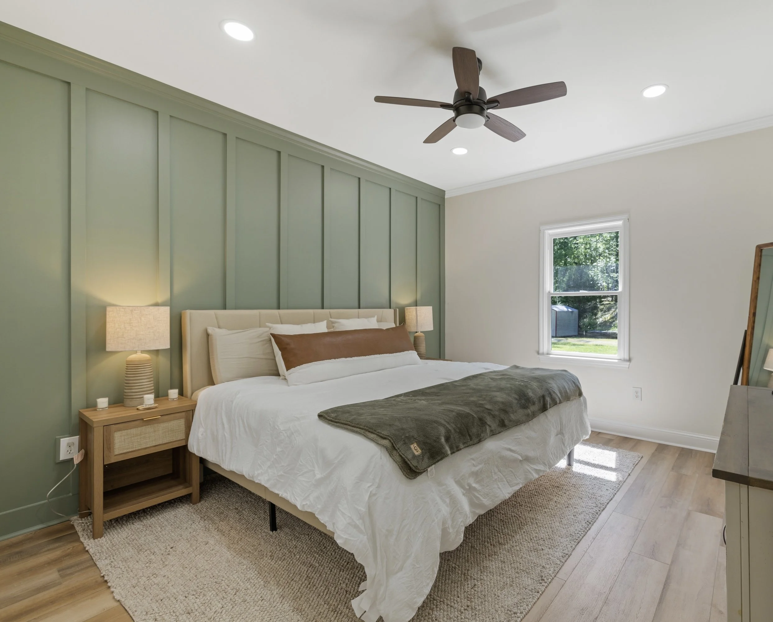A bedroom with a bed, wooden nightstands, and lamps, featuring green and white walls, hardwood flooring, a ceiling fan, and a window showing greenery outside.