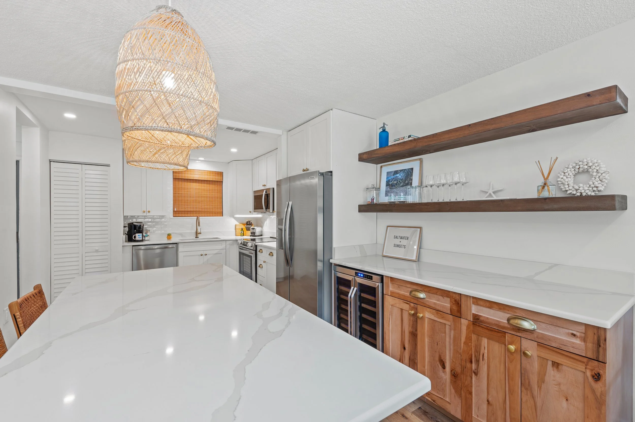 Modern coastal kitchen with white cabinets, wooden open shelves, stainless steel appliances, and a large white marble island with subtle veining. Decor includes glassware, a framed quote, and beach-themed decorations.