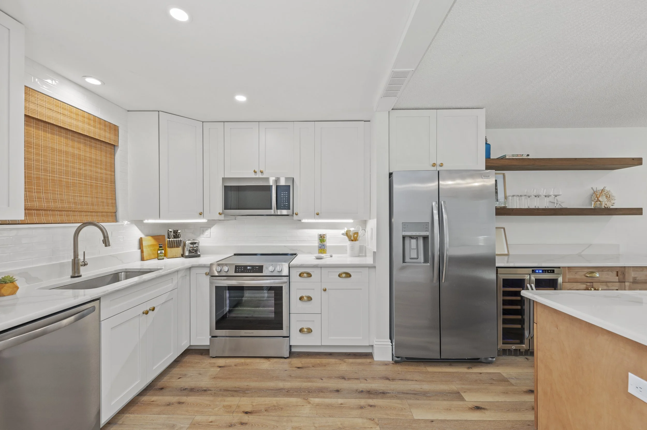 Modern kitchen with white cabinets, stainless steel appliances, wooden open shelves, and hardwood flooring.