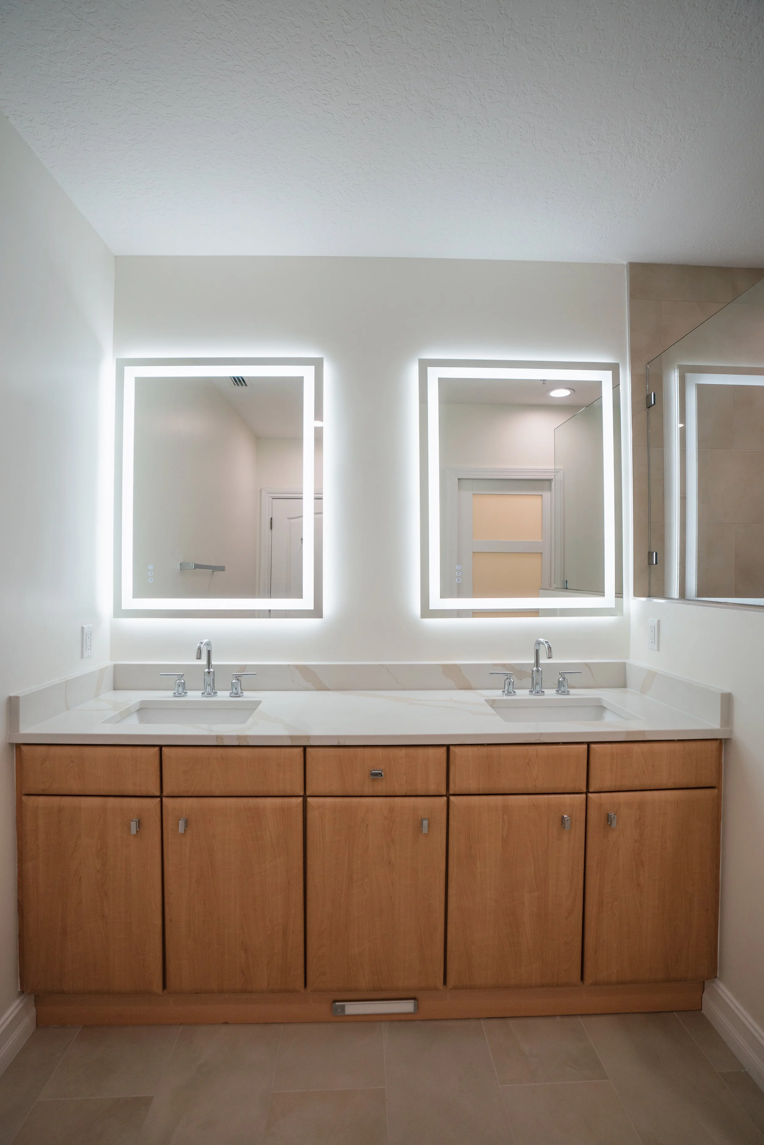 Bathroom vanity area with two illuminated rectangular mirrors, a double sink with chrome fixtures, and wooden cabinets below. A white countertop and beige tiled floor.
