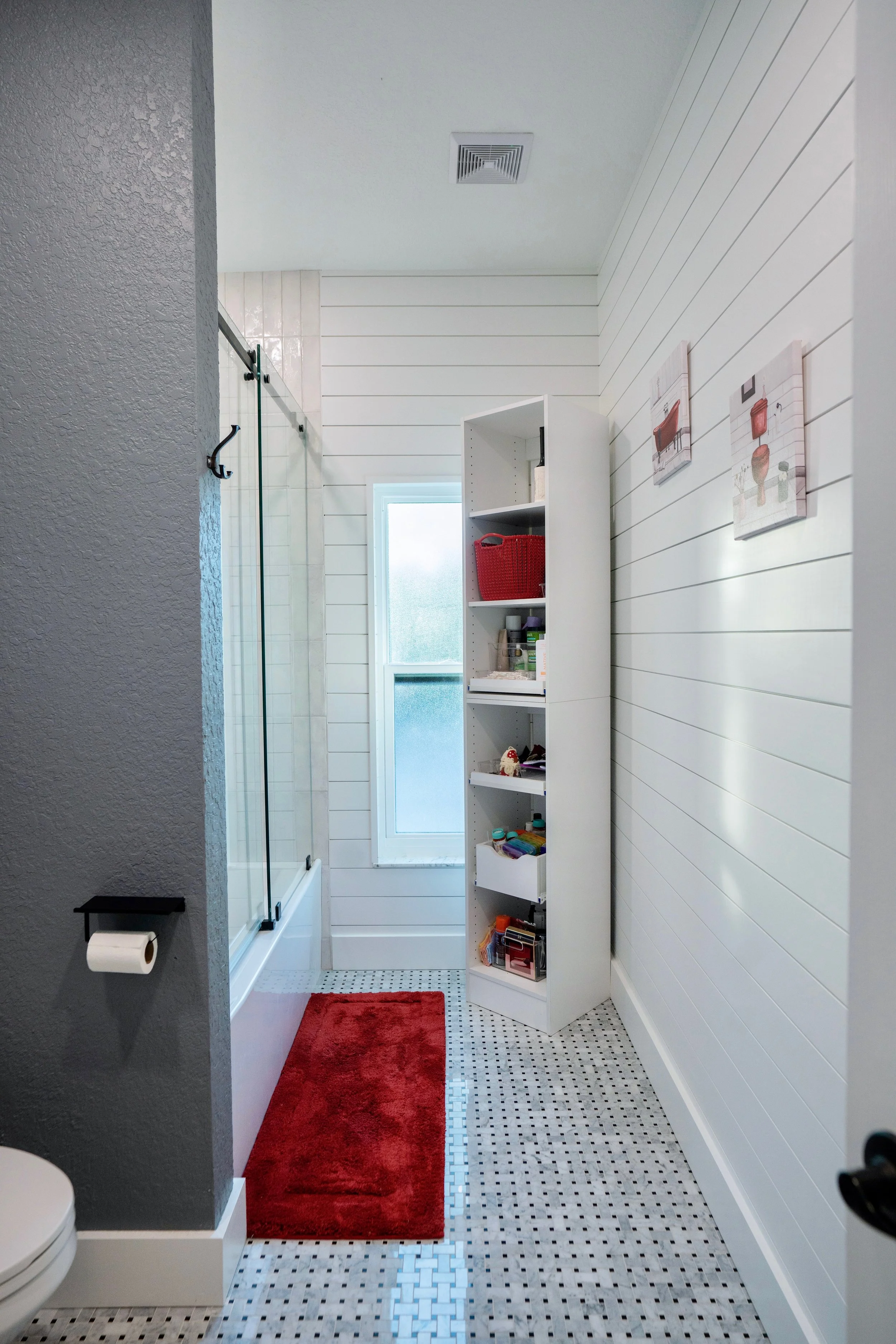 A bright bathroom with white shiplap walls, a small frosted window, a glass shower with a sliding door, a red rug on the tiled floor, and a tall white storage shelf holding various toiletries and items.