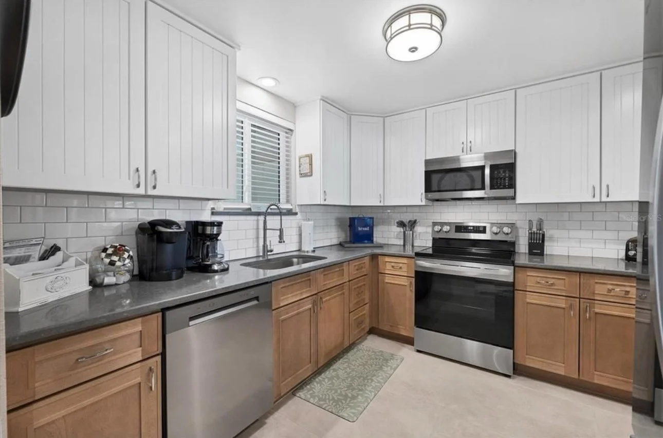 Kitchen with white upper cabinets, wooden lower cabinets, stainless steel appliances including a microwave and oven, gray countertop, white subway tile backsplash, window with blinds, small kitchen rug, and various small appliances and kitchen tools 