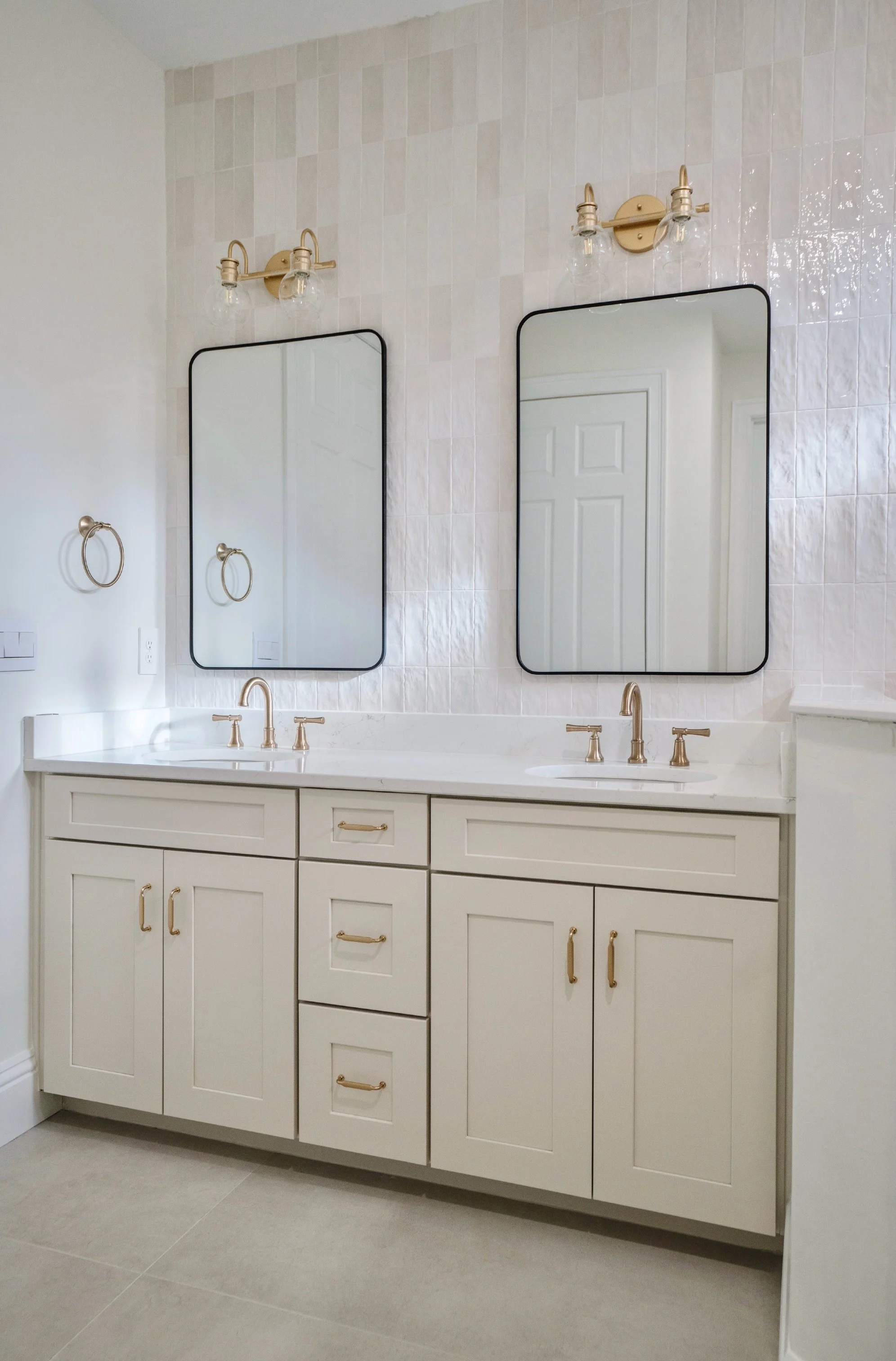 Double bathroom vanity with white cabinets, gold handles, and a white countertop with two sinks. Two rectangular mirrors with black borders are mounted above each sink, with gold light fixtures above them. There are wall-mounted gold towel rings on e