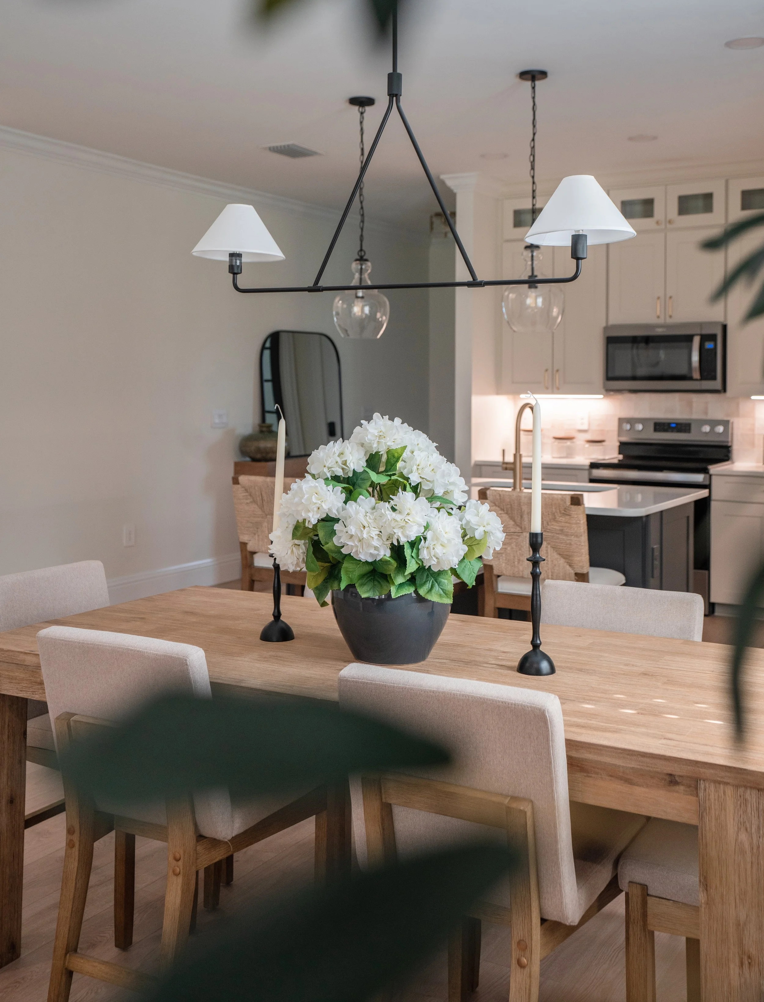 A modern dining room with a wooden table, white floral centerpiece, and beige chairs, overlooking a kitchen with white cabinets and stainless steel appliances.