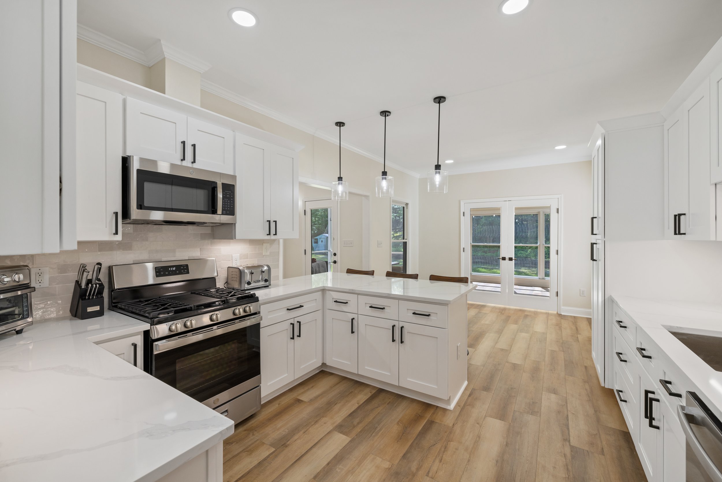 Modern kitchen with white cabinets, stainless steel appliances, light wood flooring, pendant lights, and large windows and glass doors.
