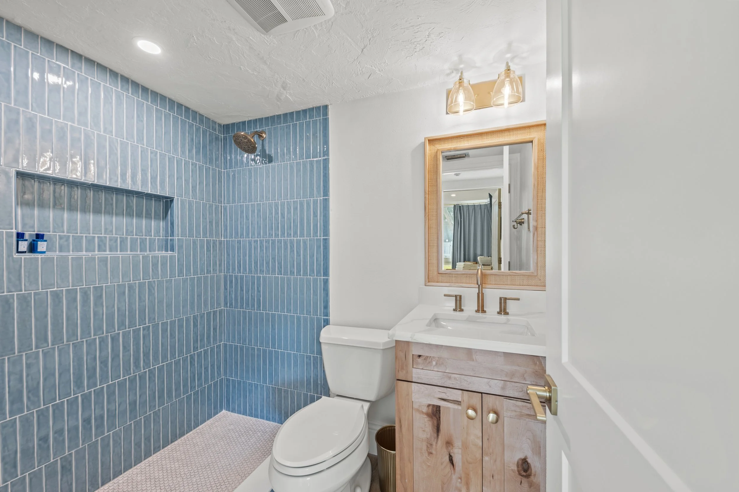 Bathroom with a walk-in shower featuring blue tiles, a white toilet, and a wooden vanity with a mirror and two light fixtures.