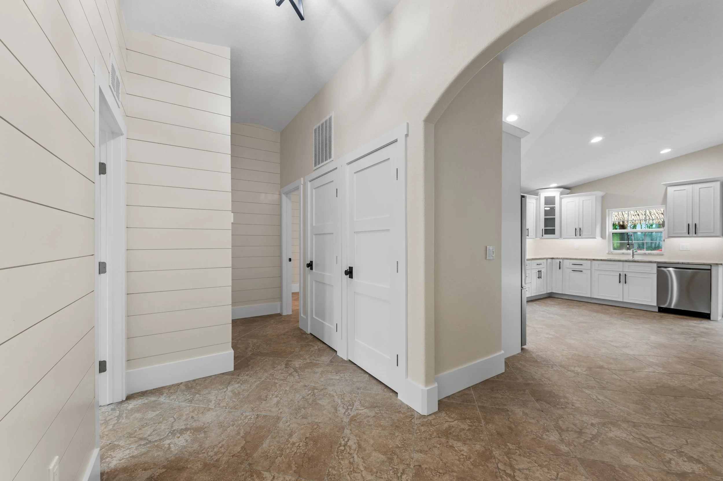 Interior view of a kitchen and hallway with white cabinets, beige walls, tiled floor, and a doorway leading to a spacious kitchen area with a window and stainless steel dishwasher.