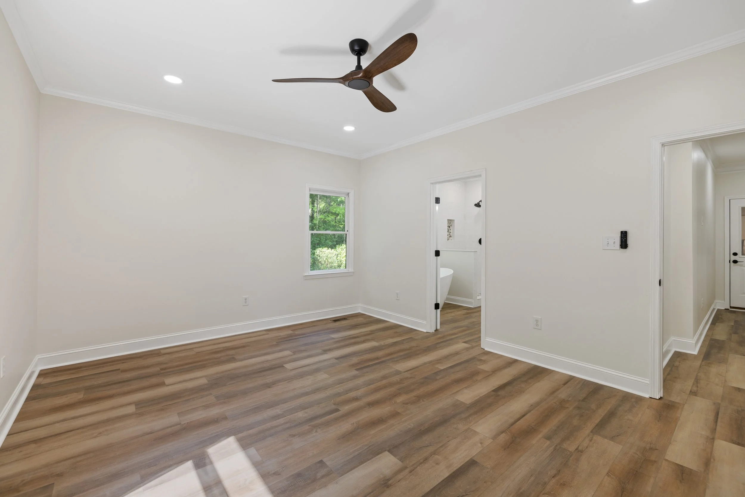 Empty room with white walls, wood flooring, ceiling fan, window, and doorway to a bathroom with bathtub.