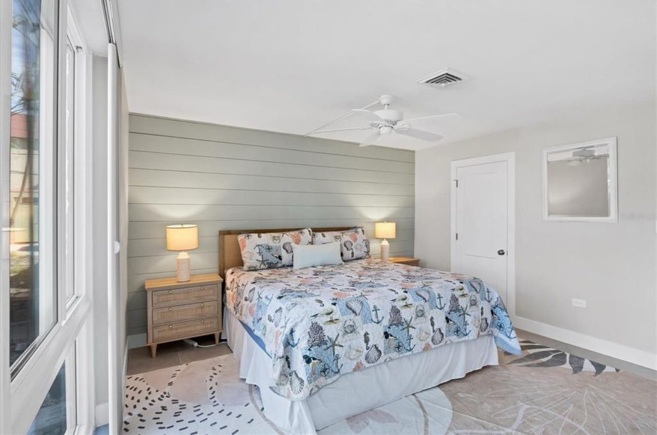 Bedroom with a large bed, ocean themed quilt, two white nightstands with lamps, located next to a window with blinds, beige area rug, gray shiplap accent wall behind the bed, white ceiling fan, white door, and window on the wall.