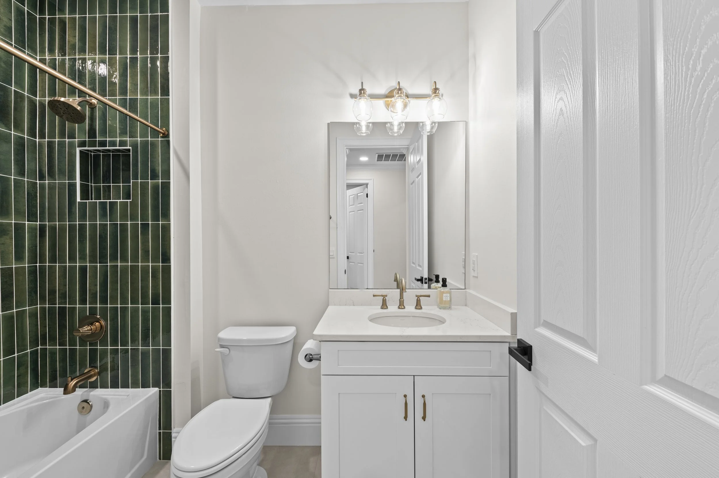 Modern bathroom with a shower and bathtub with dark green tiles, white toilet, white vanity with gold fixtures, large mirror, and a light fixture with multiple exposed bulbs.