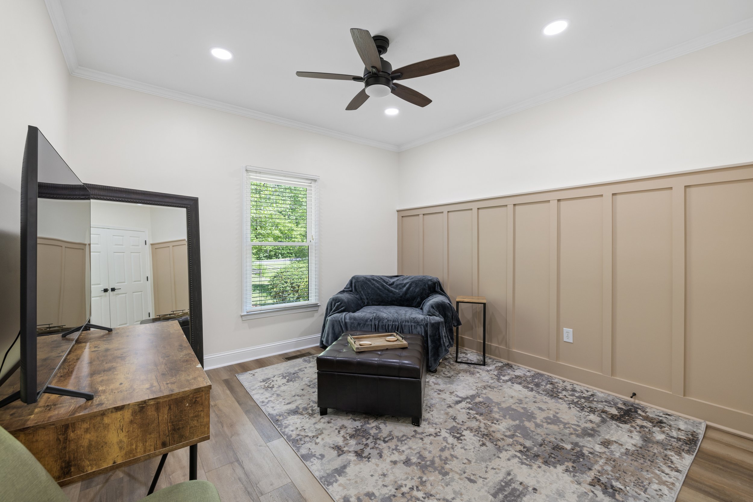 A living room with a ceiling fan, a window with green trees outside, a sofa with a blanket, a small table, a rug, and a TV on a wooden stand.