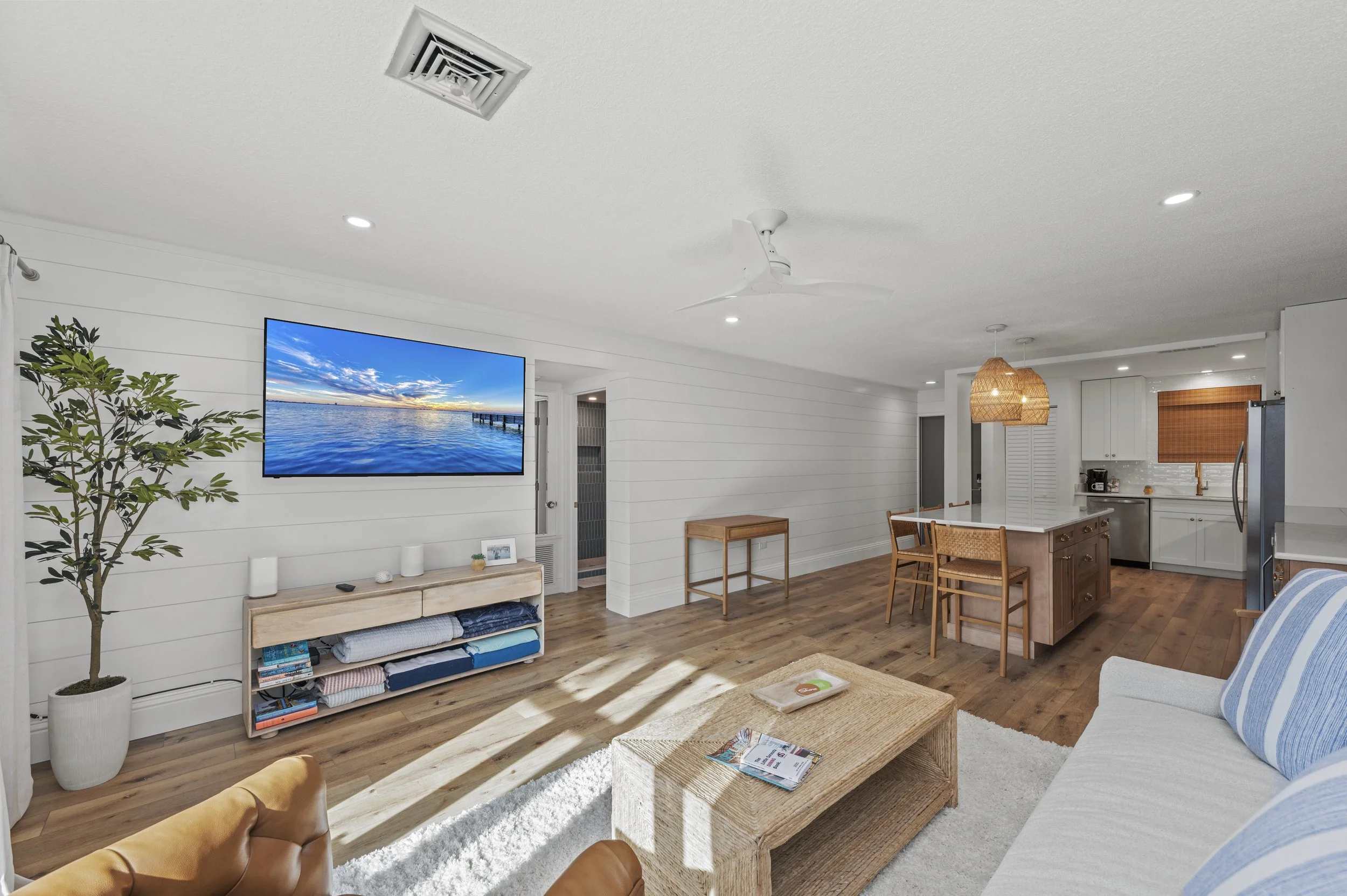 Open-plan living room and kitchen with white shiplap walls, hardwood floors, a large flat-screen TV, a potted plant, a wooden media console, a beige couch with blue and white striped pillows, a rattan coffee table, a white ceiling fan, a kitchen isla