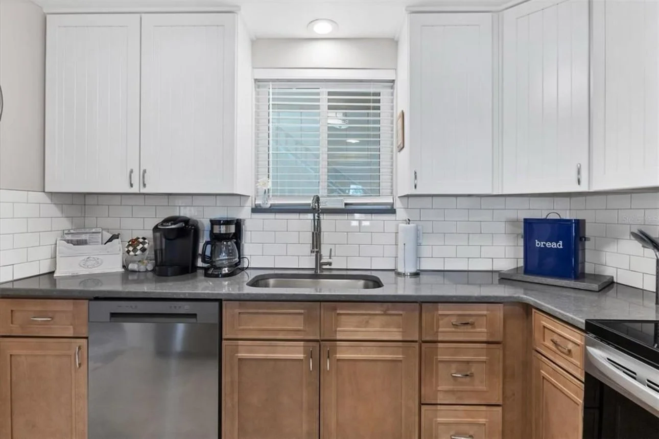 Kitchen with brown cabinets, white upper cabinets, a window above the sink, white subway tile backsplash, and various small appliances including a coffee maker, a coffee pot, and a bread bag on the countertop.