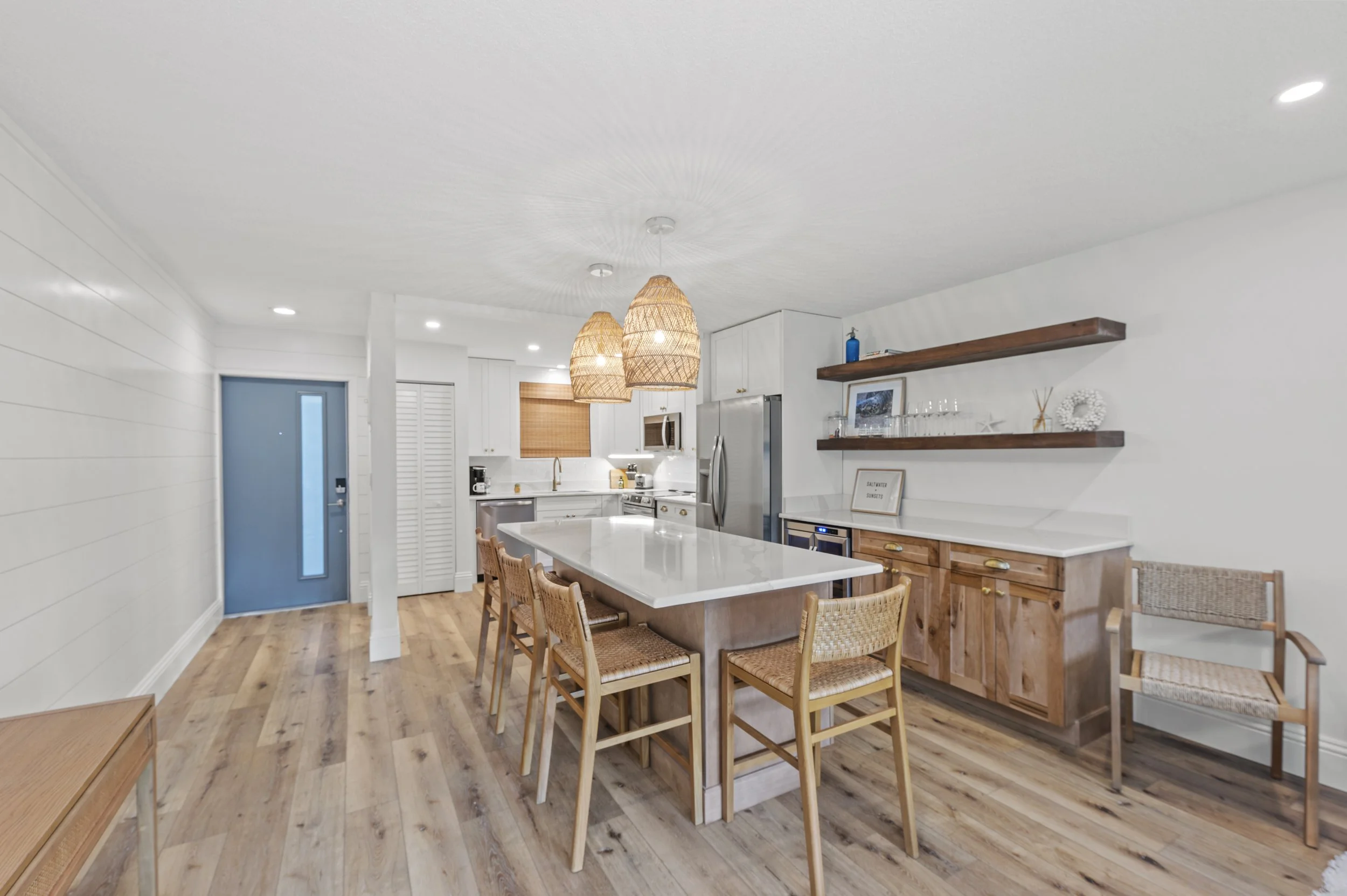 Bright kitchen with white cabinetry, a large island with a white countertop, wooden stools, and open wooden shelves with decorative items, under woven pendant lights, with light hardwood floors.