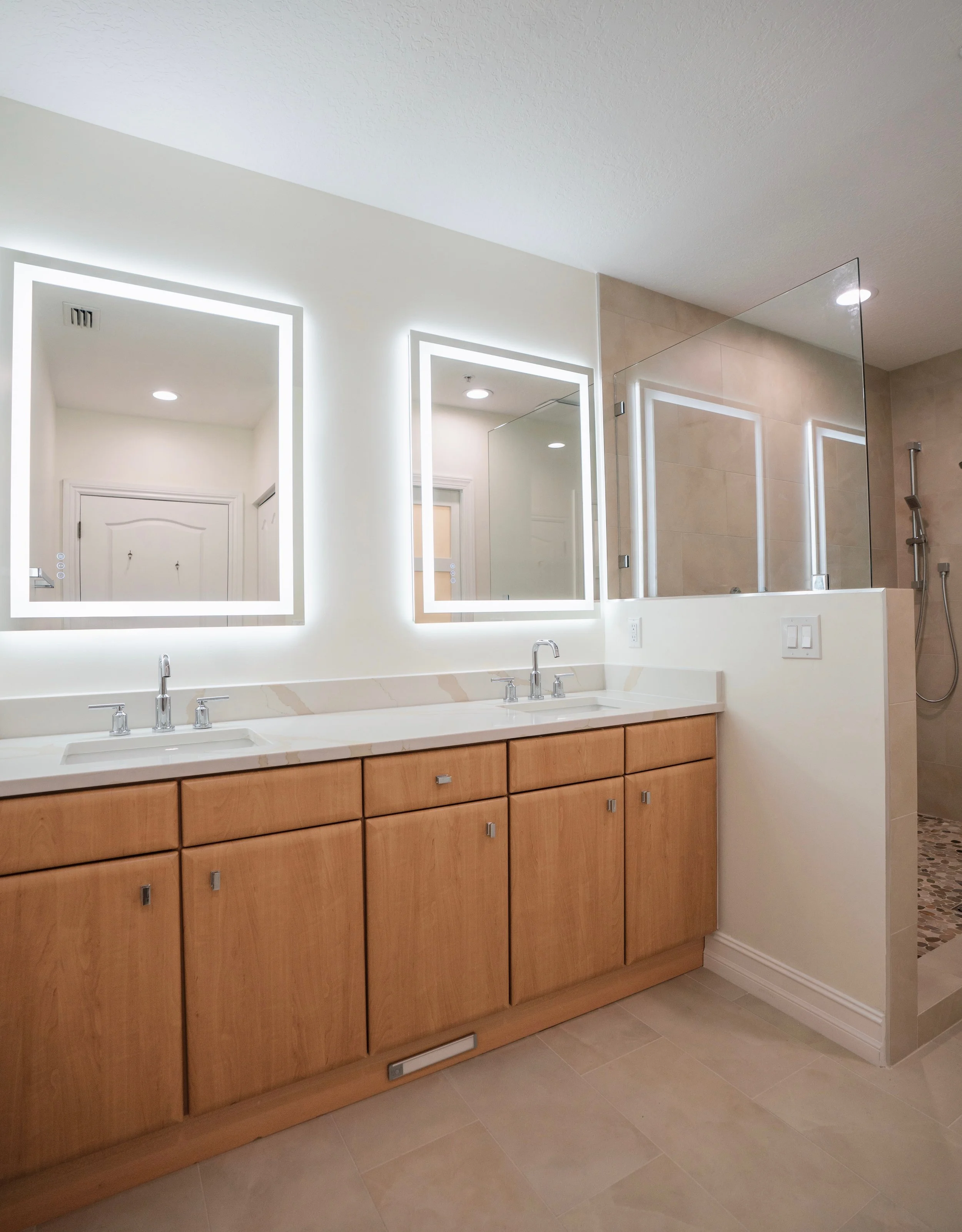 Bathroom with double vanity, illuminated mirrors, wood cabinets, beige floor tiles, and a glass-enclosed shower with tan tiles.