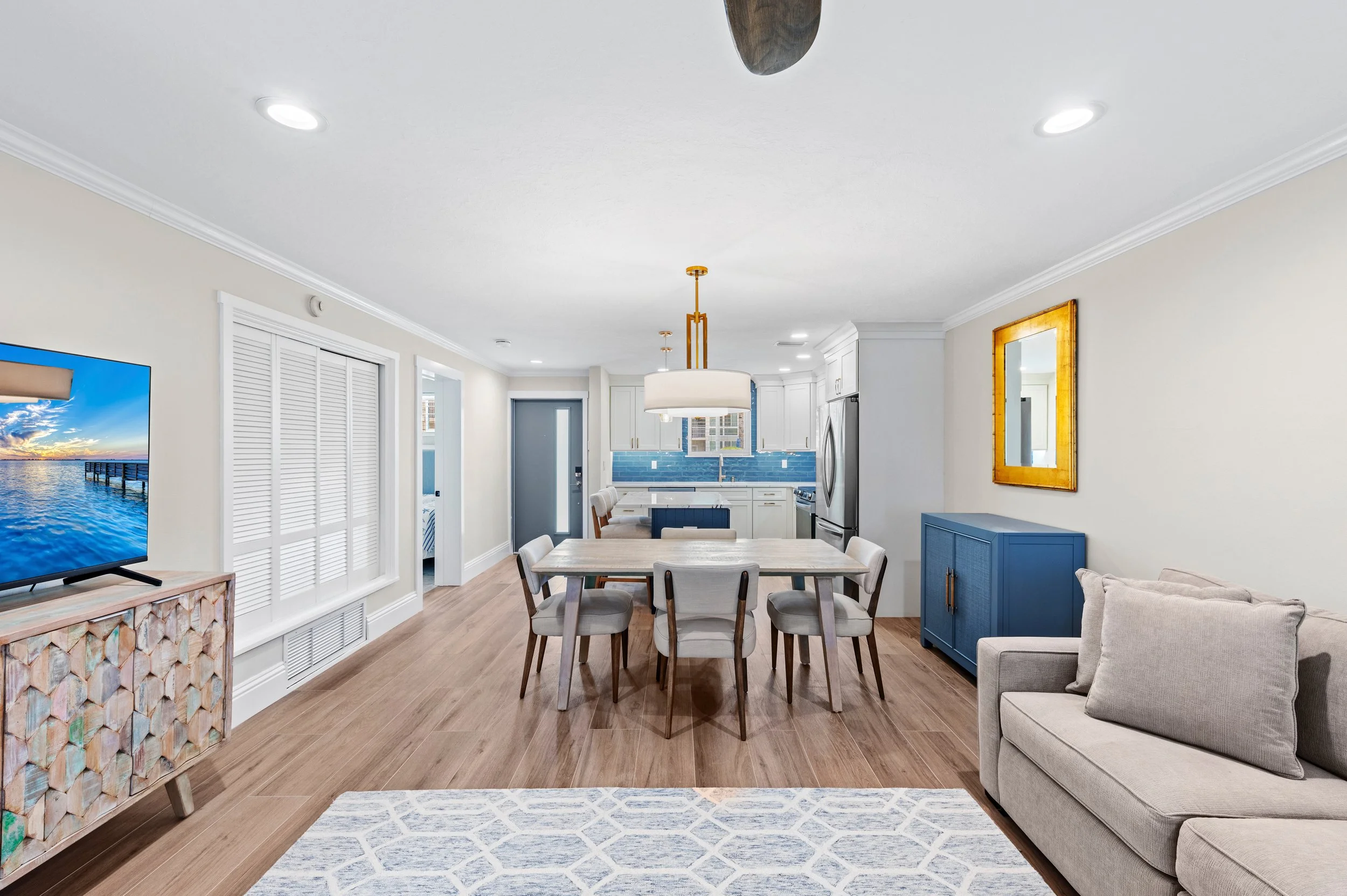 Open-concept living and dining area with kitchen in the background, featuring light-colored walls, hardwood floors, a gray sofa, a blue sideboard, a dining table with eight chairs, ceiling lights, and a television on a decorative cabinet.