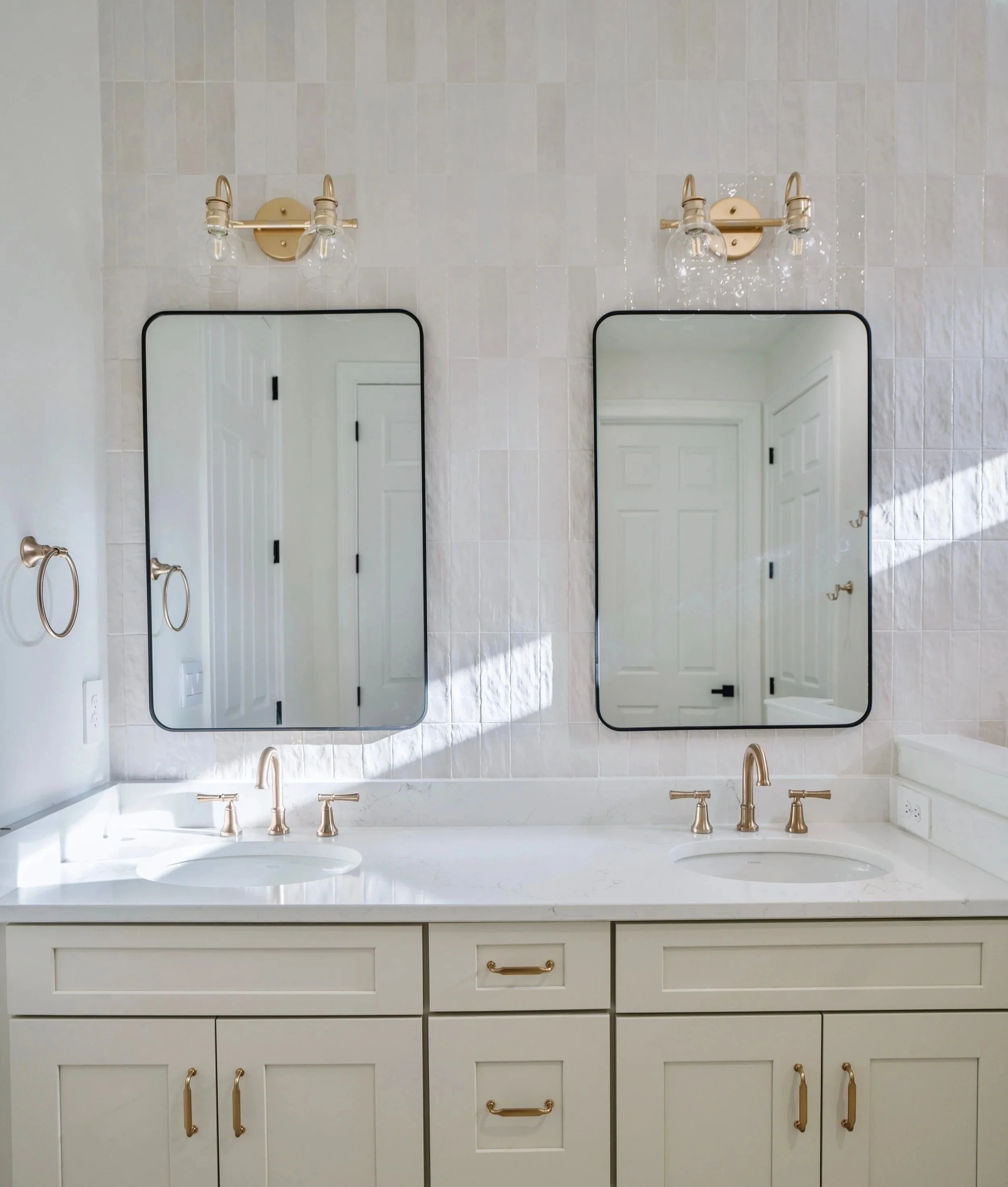 Double bathroom vanity with two oval mirrors, brass faucets, two wall-mounted light fixtures, and beige cabinetry with gold handles, white tiled wall.