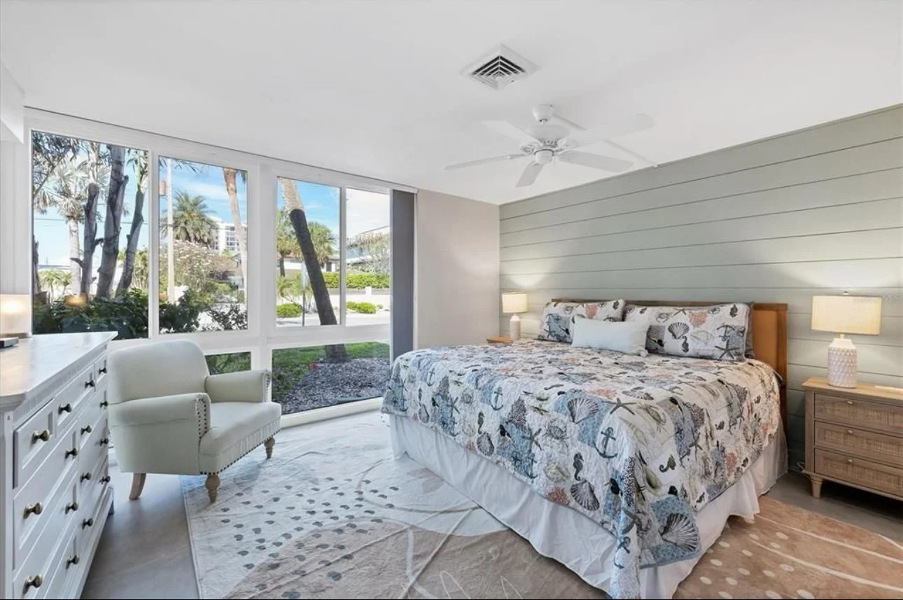 A bedroom with a large bed, ocean-themed bedding, two nightstands with lamps, a white armchair, and large windows showing palm trees outside.