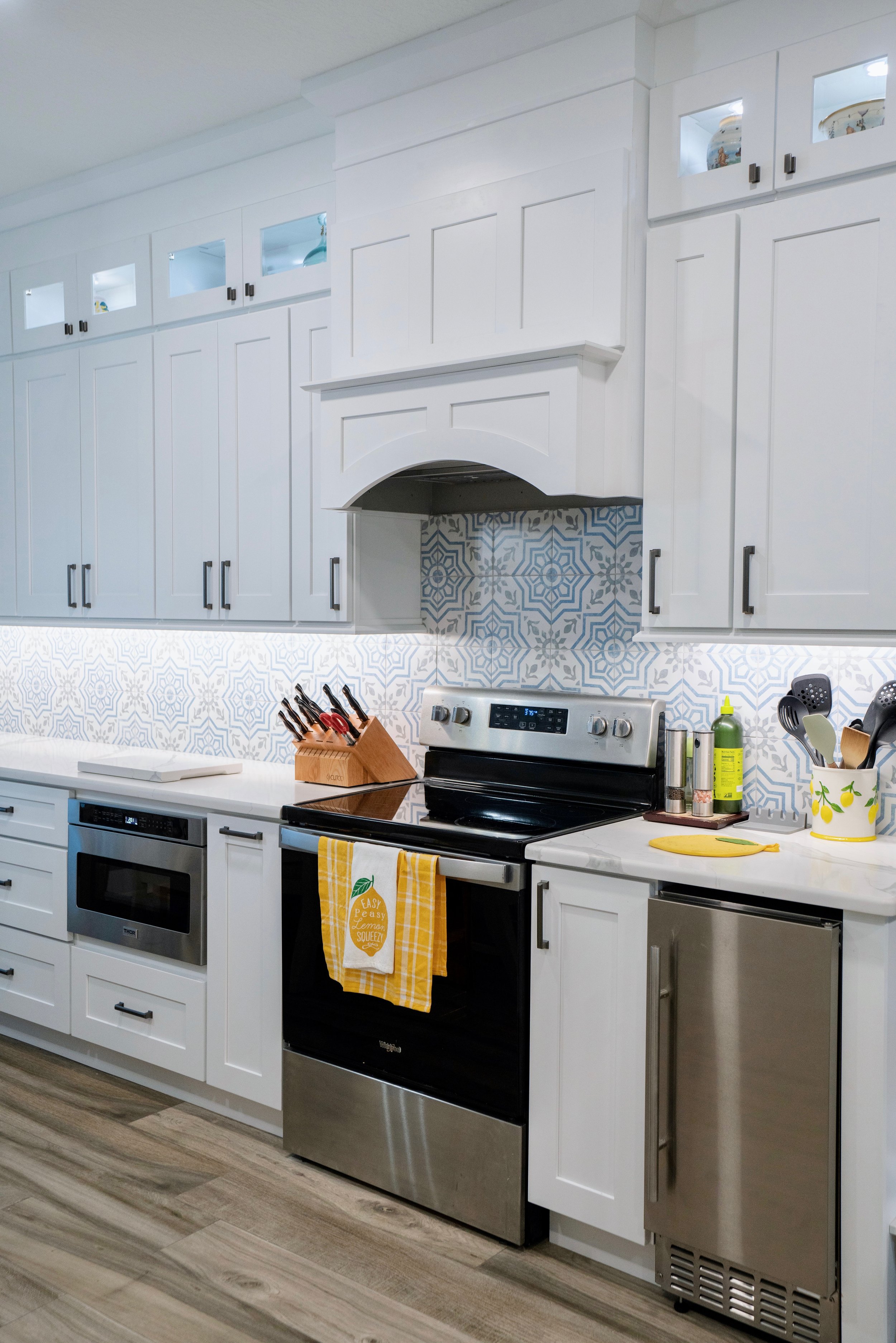 Modern white kitchen with patterned blue and white backsplash, stainless steel appliances including oven and mini-fridge, black handles on cabinets, and various kitchen utensils on the counter.