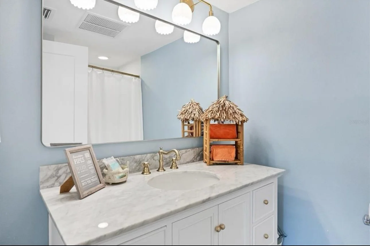 Bathroom vanity with white cabinets and marble countertop, a large mirror, and decorative items including a framed sign, a small box, and a wooden shelf with two thatched-roof huts holding orange towels.