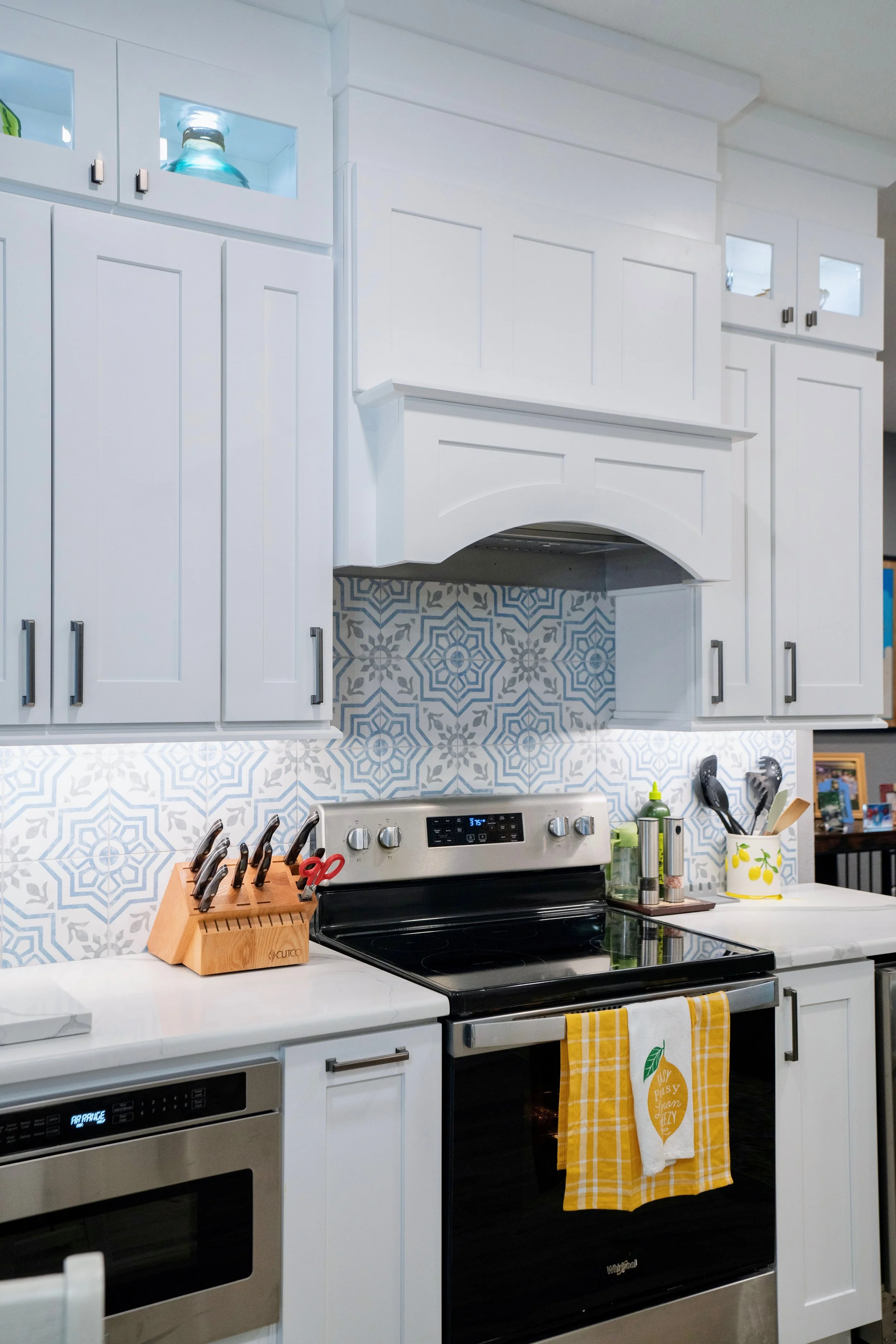 Modern kitchen with white cabinets, black stove, and patterned blue and white backsplash. Contains knives in a wooden block, scissors, and utensils on the counter.